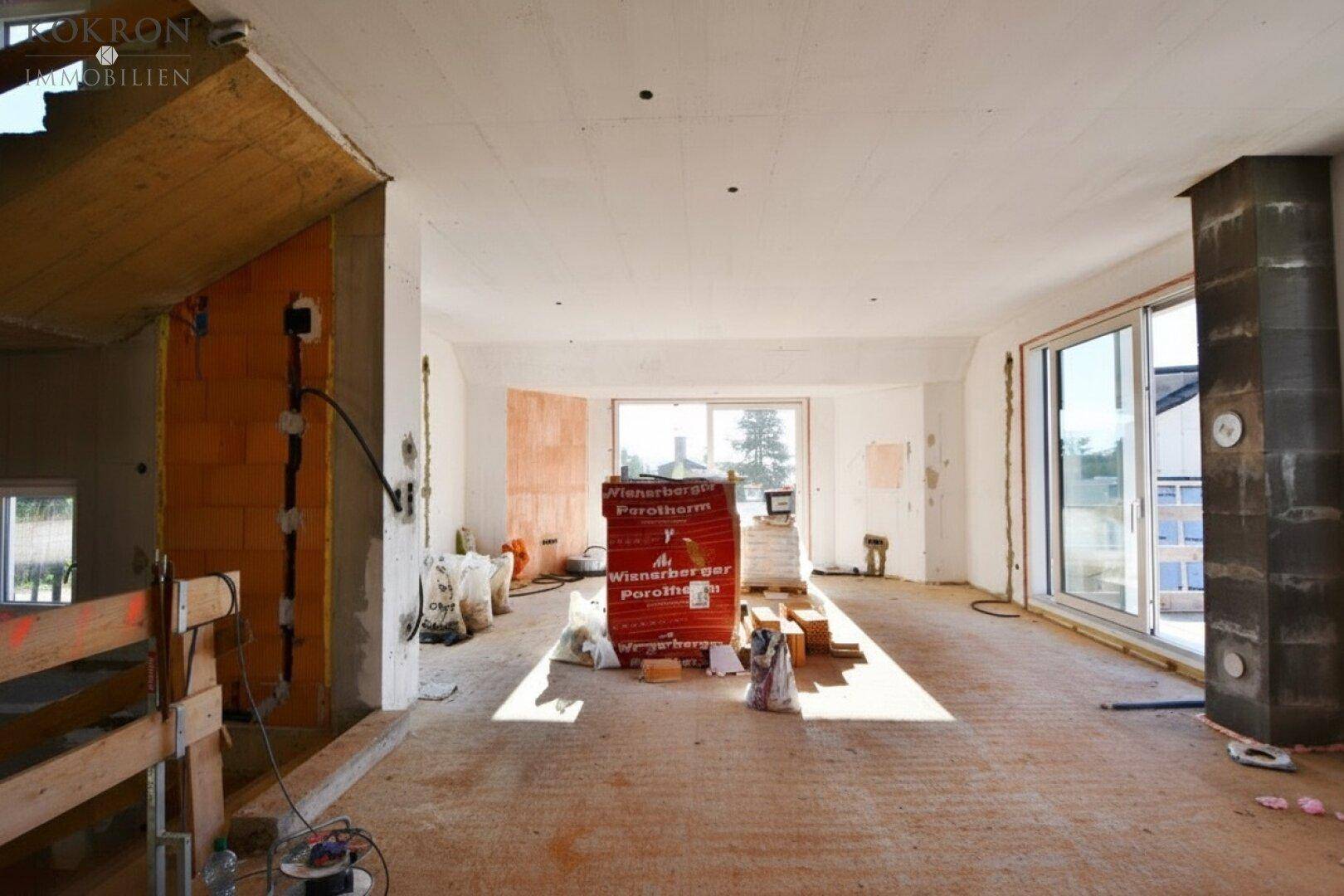 Großer offener Raum im Rohbau mit Baumaterialien und viel Tageslicht durch Fenster.