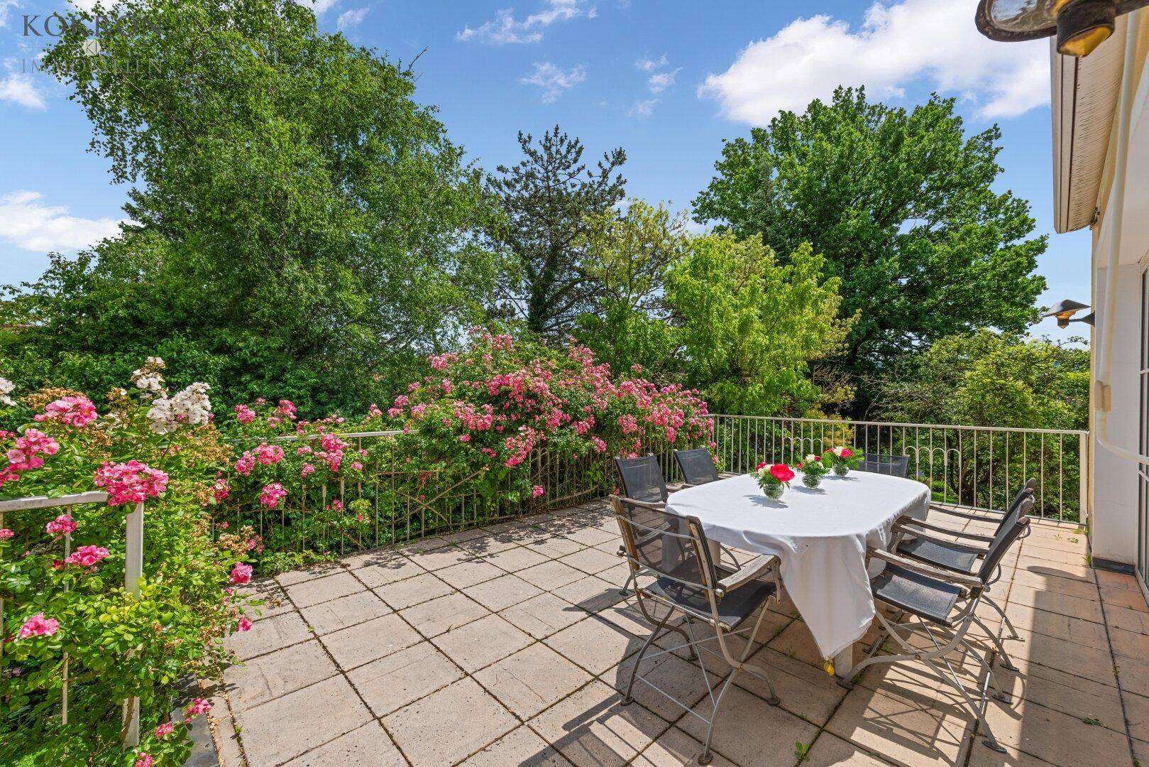 Sonniger Terrassenbereich mit Esstisch und Stühlen, umgeben von blühenden Rosen und viel Grün.