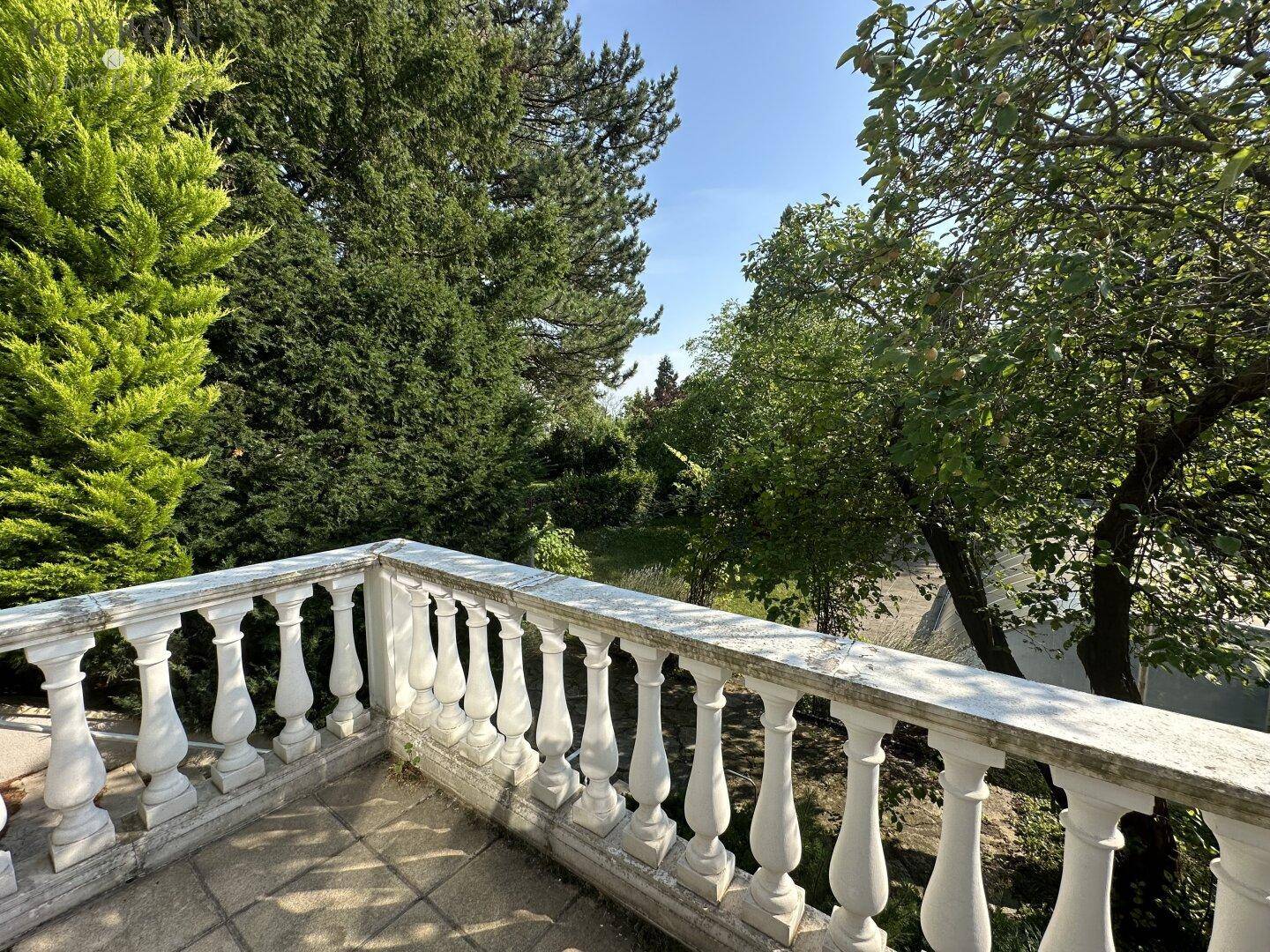 Terrasse mit klassischer Balustrade und Fliesenboden, umgeben von üppigem Grün und Bäumen.