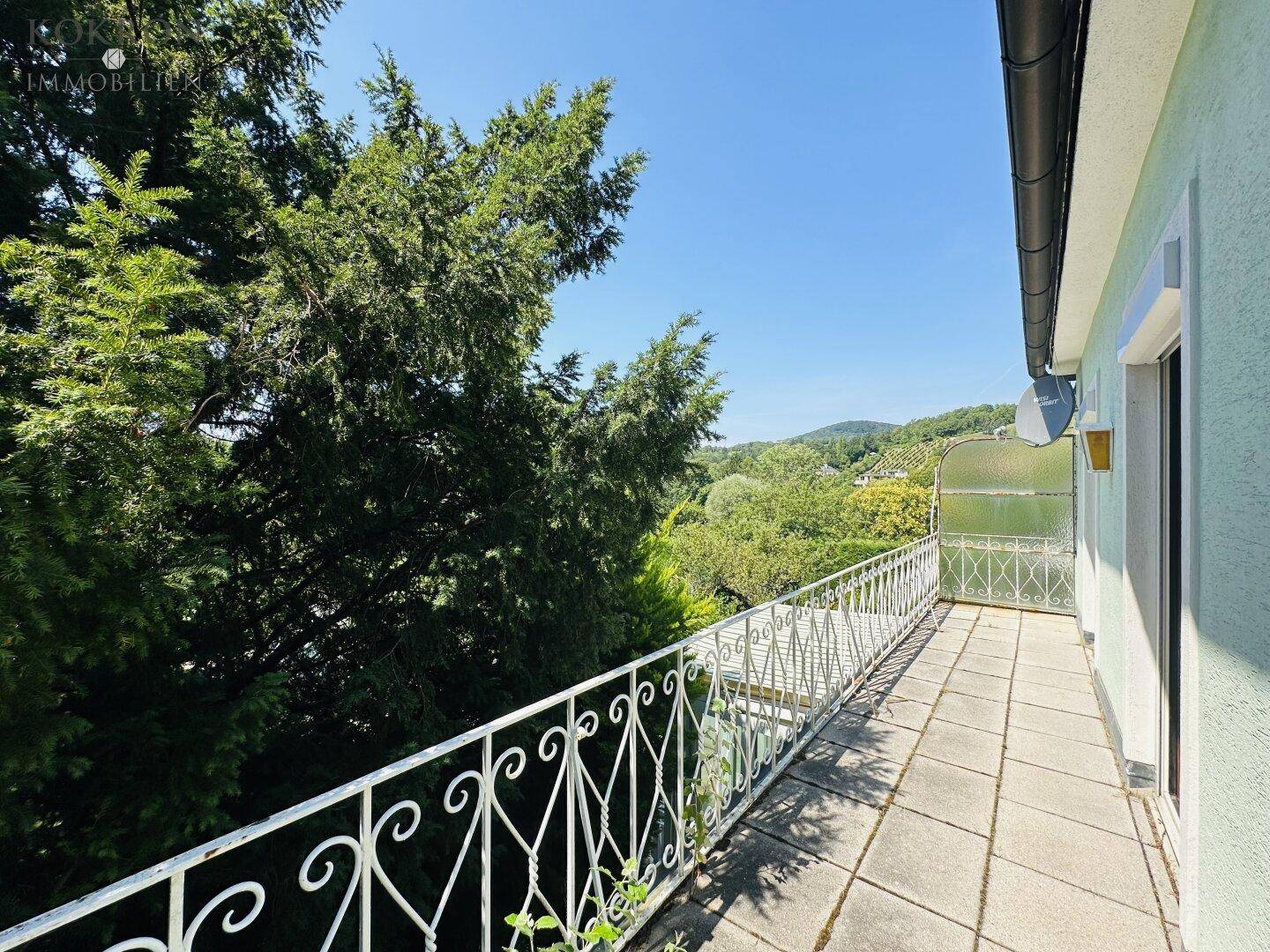 Langer Balkon mit Metallgeländer und Fliesenboden, bietet einen weiten Blick ins Grüne und auf Hügel.