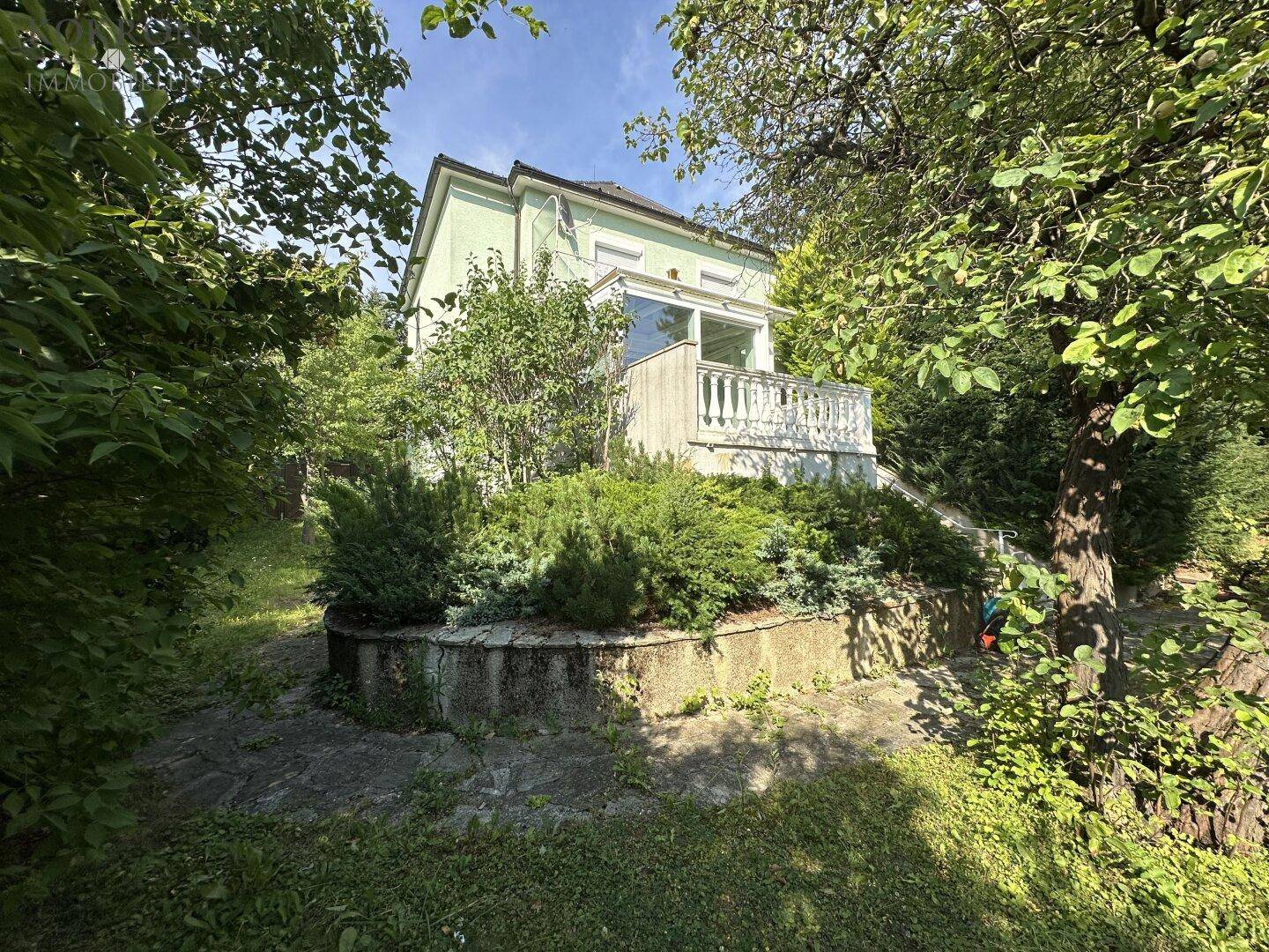 Grünes Haus umgeben von üppiger Vegetation und Bäumen mit einem Balkon im oberen Bereich.