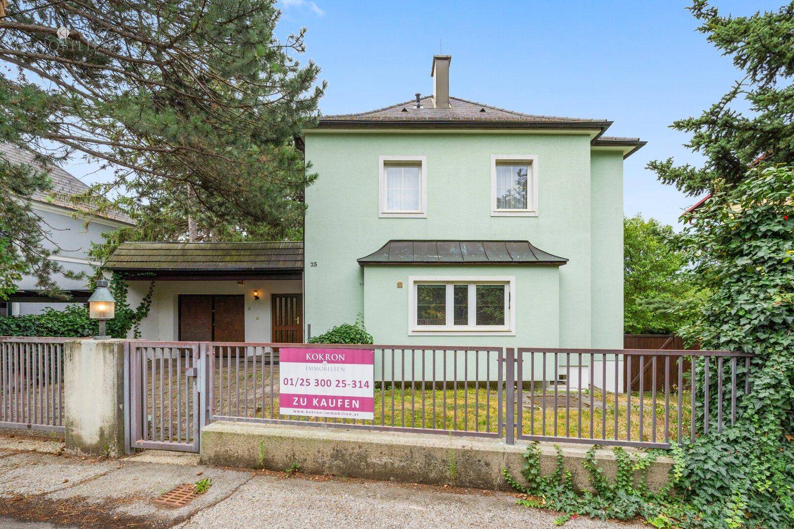 Vorderansicht des hellgrünen Hauses mit Zaun, Einfahrt und einem Verkaufsschild im Vorgarten.
