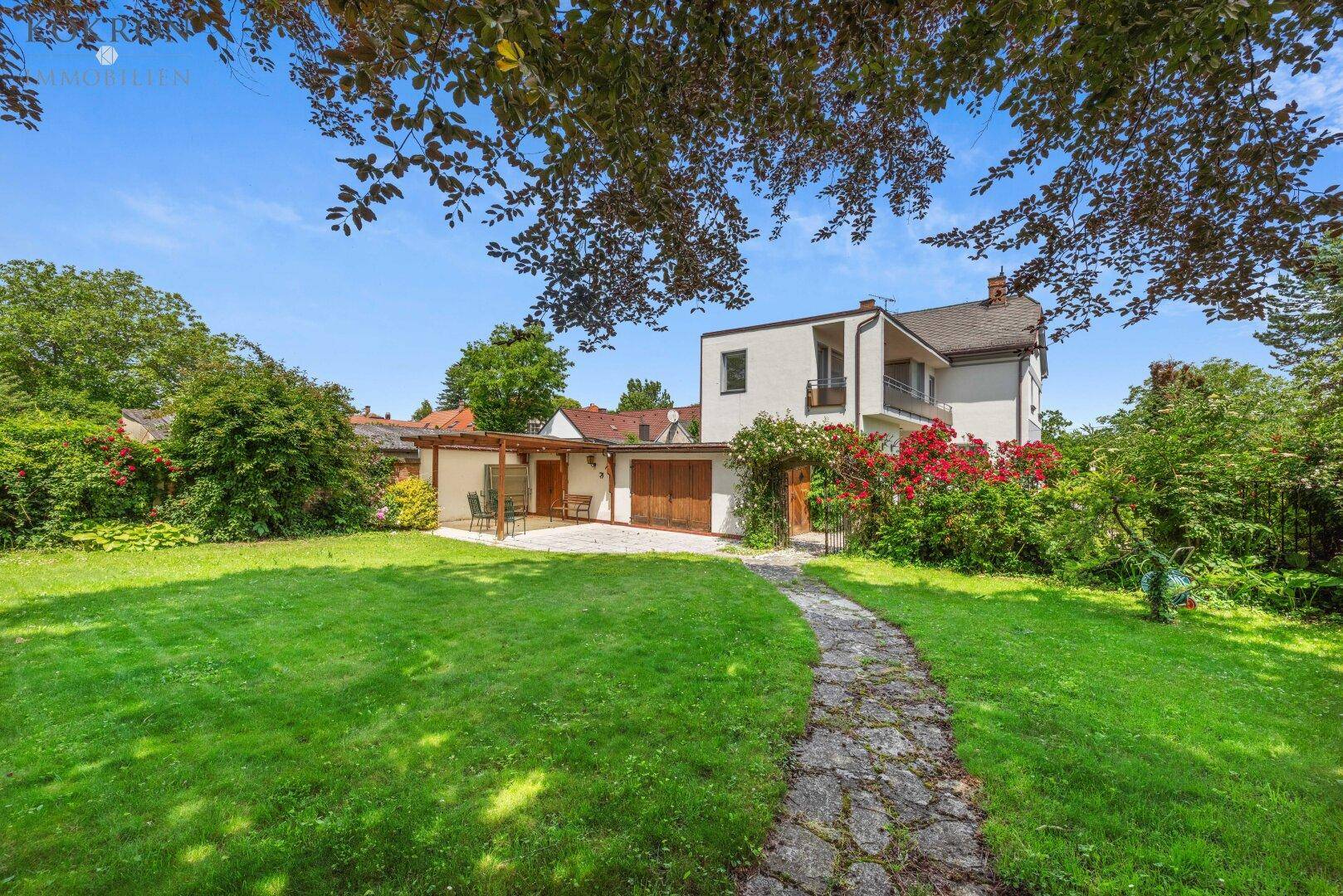 Blick auf die Rückseite des Hauses mit einer Terrasse und einem großen, gepflegten Garten mit Rasenfläche und Bäumen.