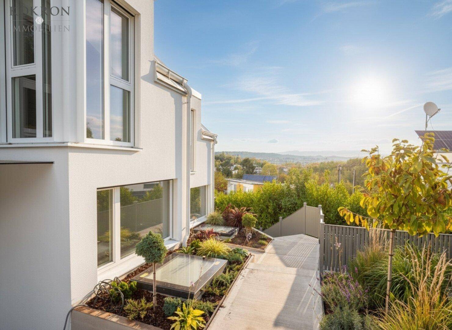 Moderne Hausfassade mit großen Fenstern und einer sonnigen, bepflanzten Außenanlage.