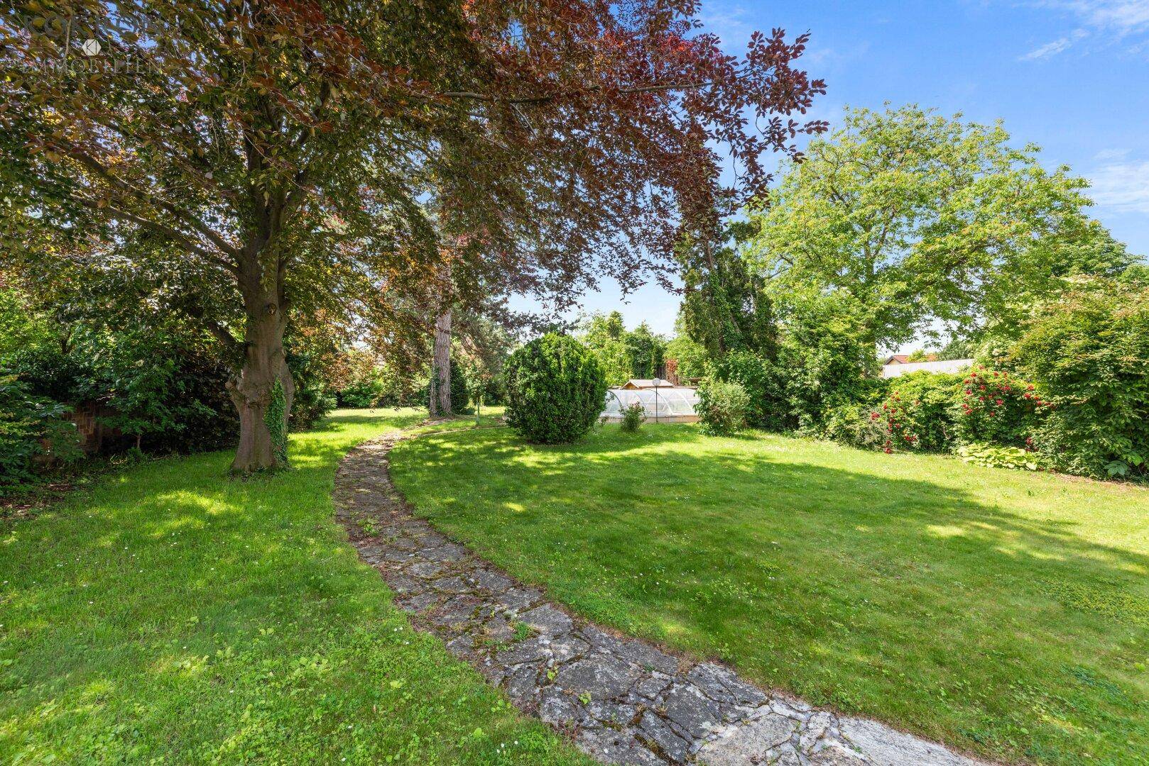 Ein weitläufiger grüner Garten mit altem Baumbestand und einem charmanten Steinweg, der durch die Rasenfläche führt.