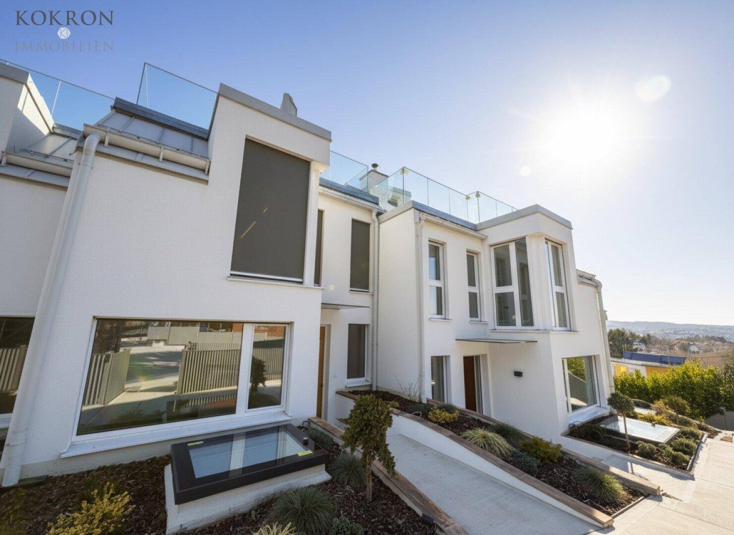 Moderne Architektur mit weißer Fassade, großen Fenstern und Dachterrasse unter strahlendem Himmel.