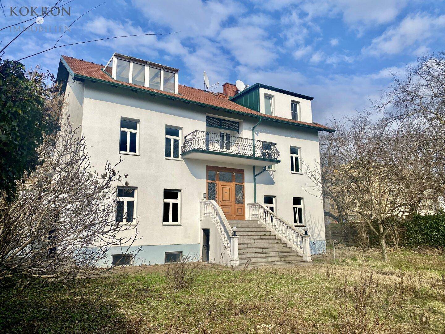 Rückansicht des Gebäudes mit Außentreppe, Balkon und Dachgauben unter blauem Himmel.