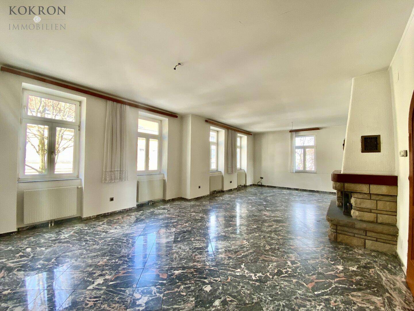 Helles Wohnzimmer mit großen Fenstern, dunklem Marmorboden und traditionellem Kamin.
