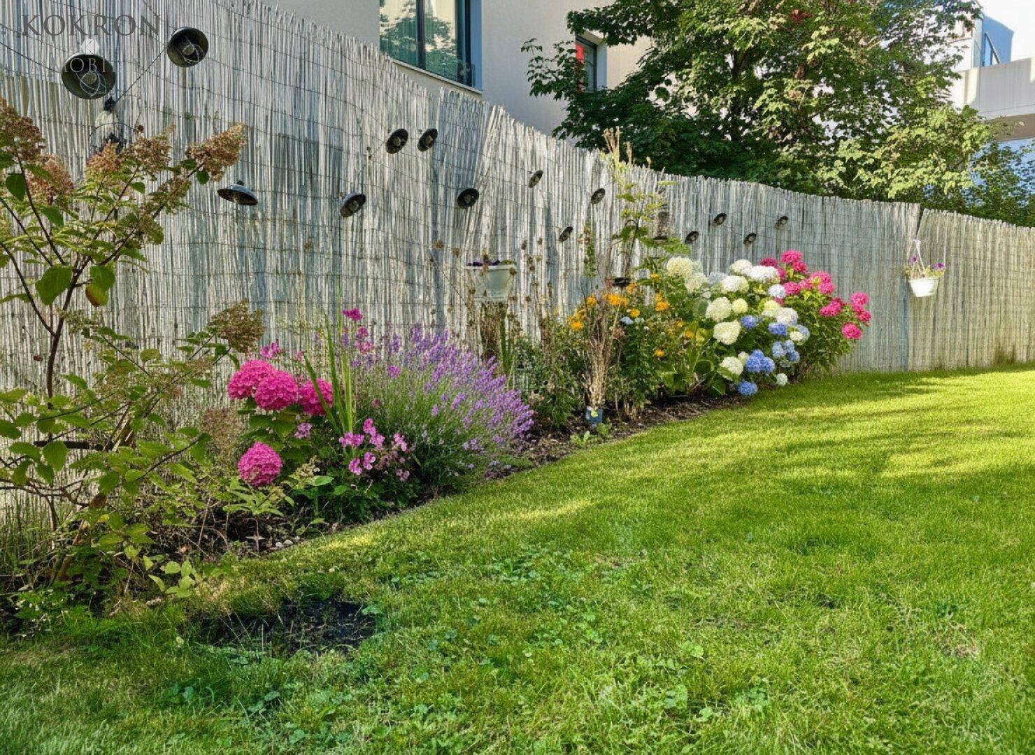 Gepflegter Gartenbereich mit blühenden Blumenbeeten, grünem Rasen und einem Sichtschutzzaun.