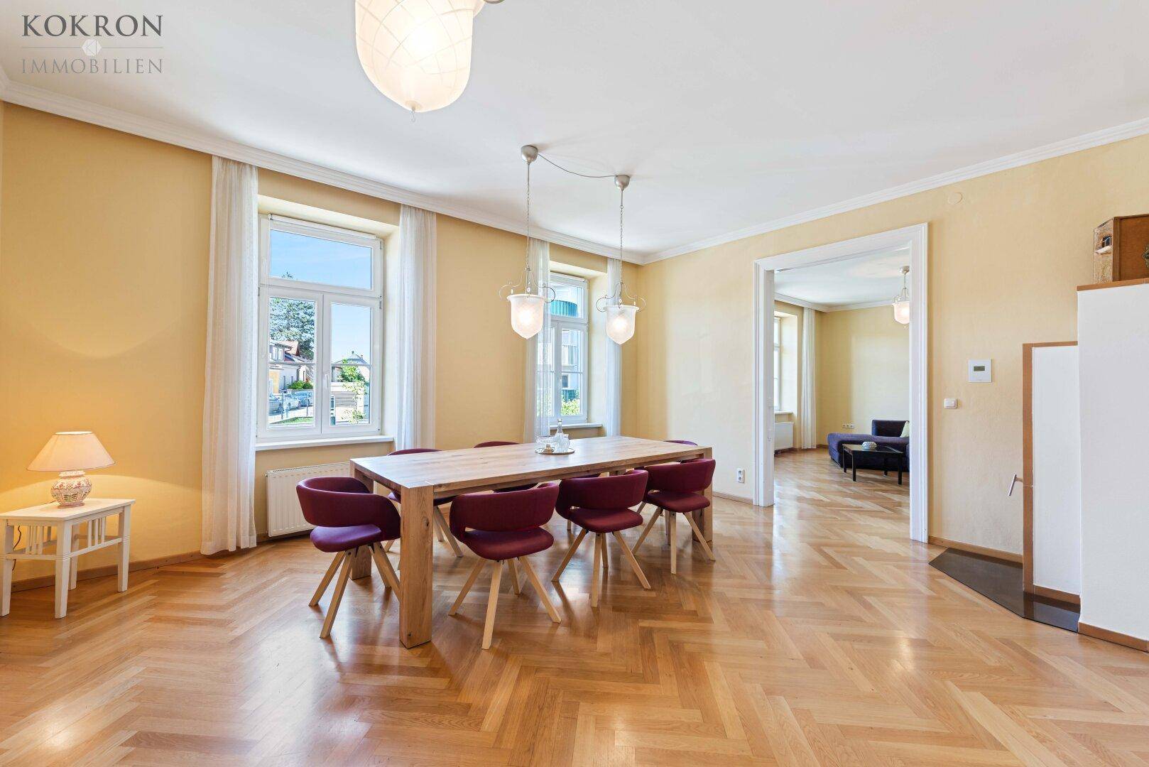 Geräumiges Esszimmer mit Fischgrätparkett, hohen Fenstern und Zugang zu einem weiteren Raum.