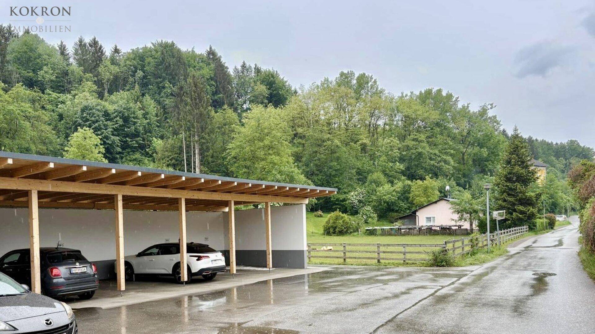 Überdachter Carport mit mehreren Stellplätzen neben einer Straße und bewaldeter Umgebung.