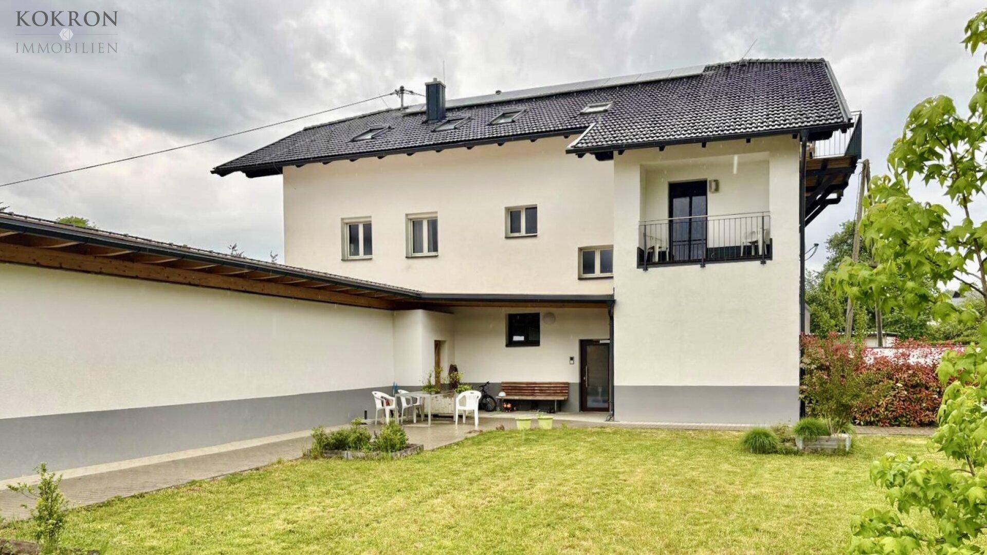 Modernes Wohnhaus mit weißer Fassade, dunklem Dach und kleinem Balkon, umgeben von Grünflächen.