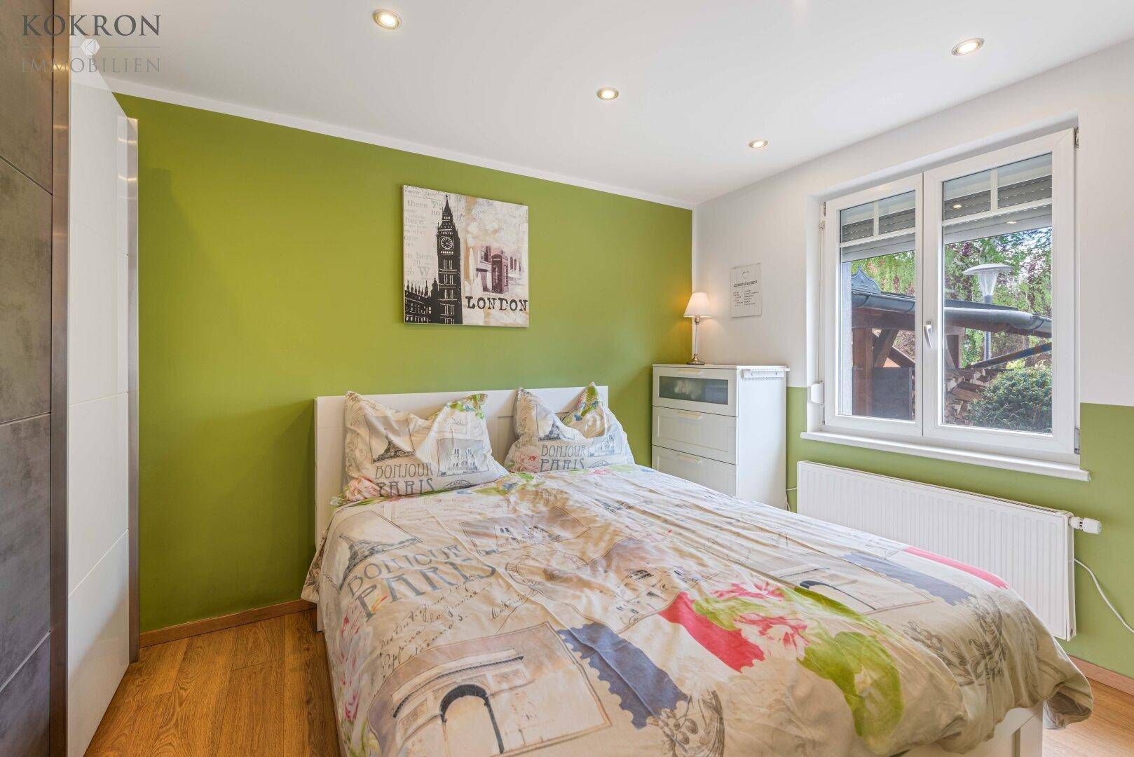 Gemütliches Schlafzimmer mit grüner Akzentwand, Holzboden und großem Fenster für viel Licht.