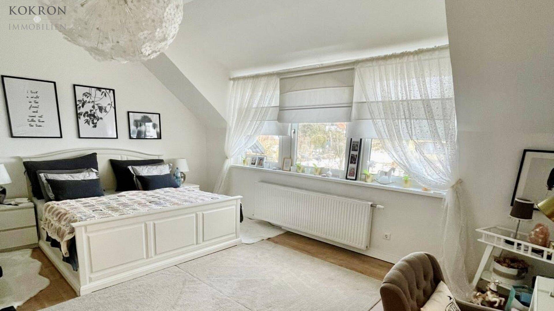 Helles Schlafzimmer mit weißem Bett, Dachschrägen und zwei Fenstern mit Vorhängen.