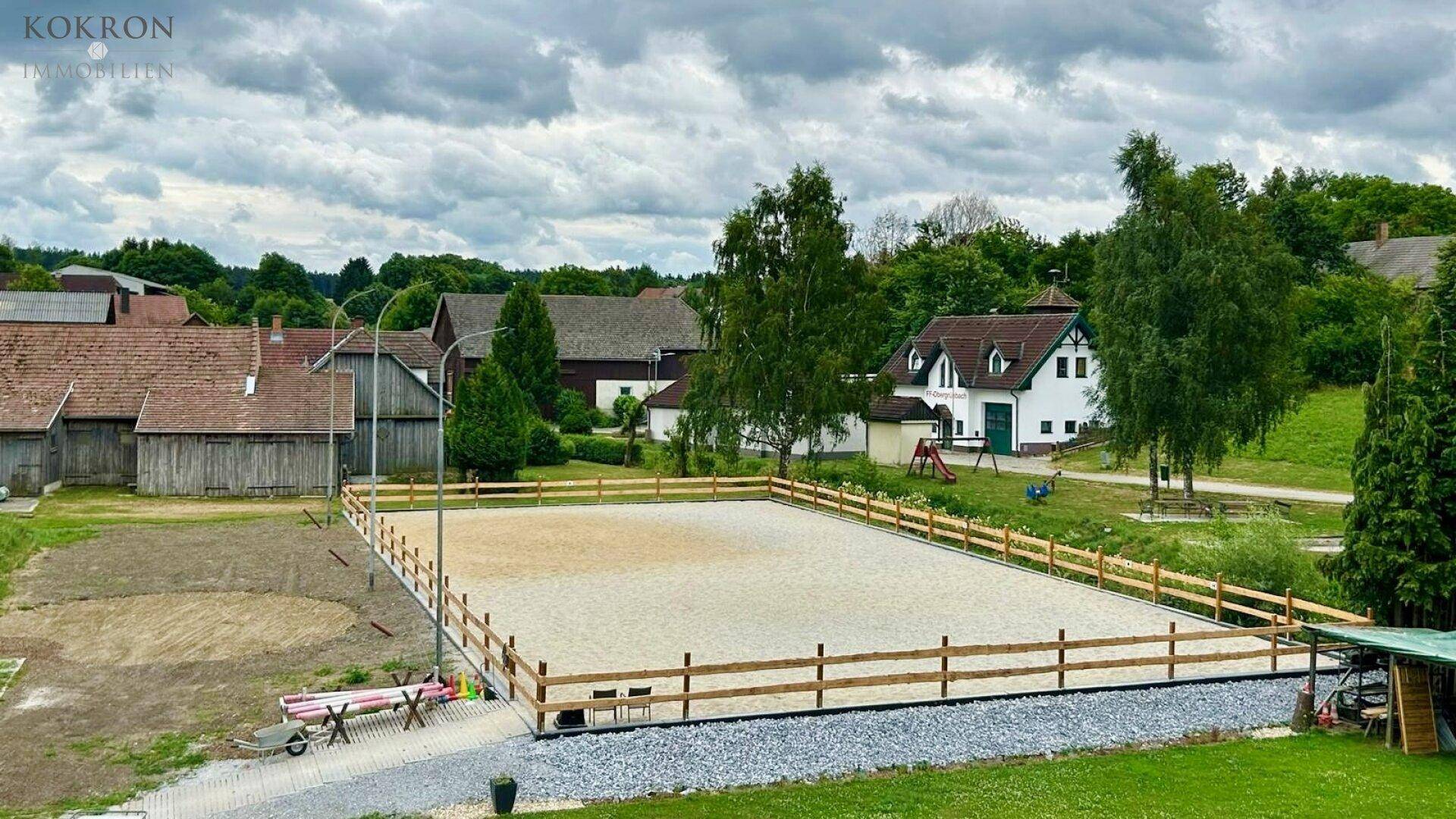 Reitplatz mit Holzzaun und ländlicher Umgebung, im Hintergrund ein Wohnhaus mit Spielplatz.