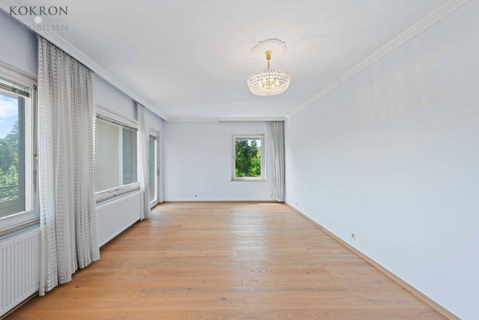 Heller, leerer Raum mit Holzboden, großen Fenstern und einem eleganten Kronleuchter an der Decke.