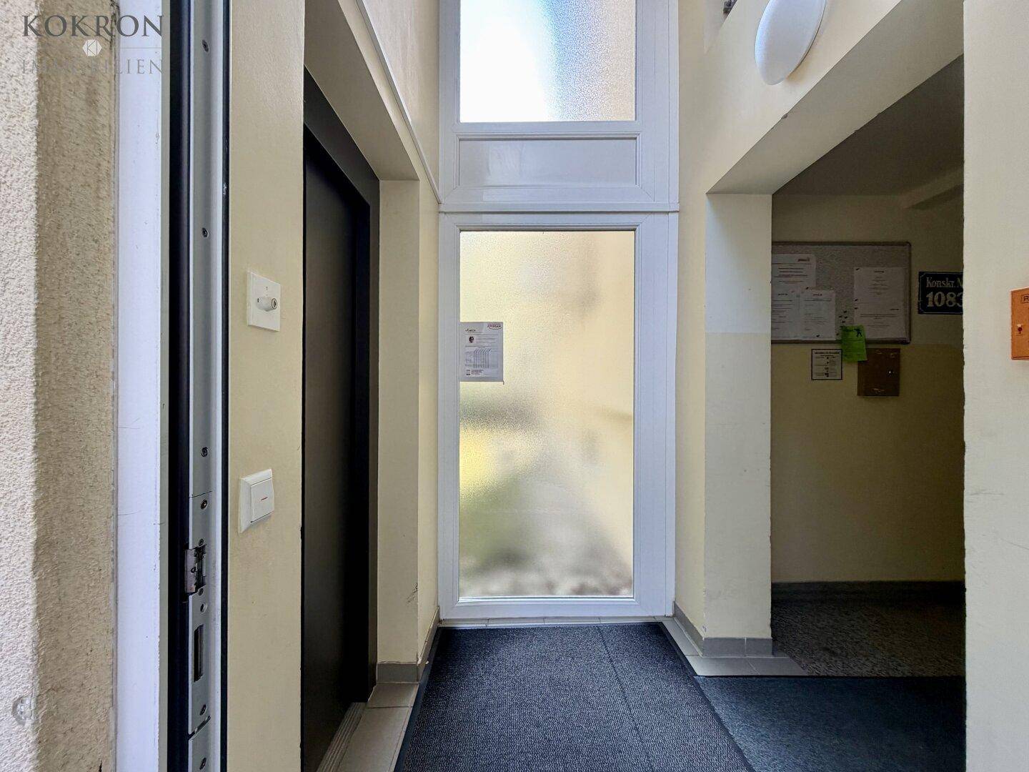 Eingangsbereich mit Aufzug, heller Wandfarbe und einer undurchsichtigen Glastür.