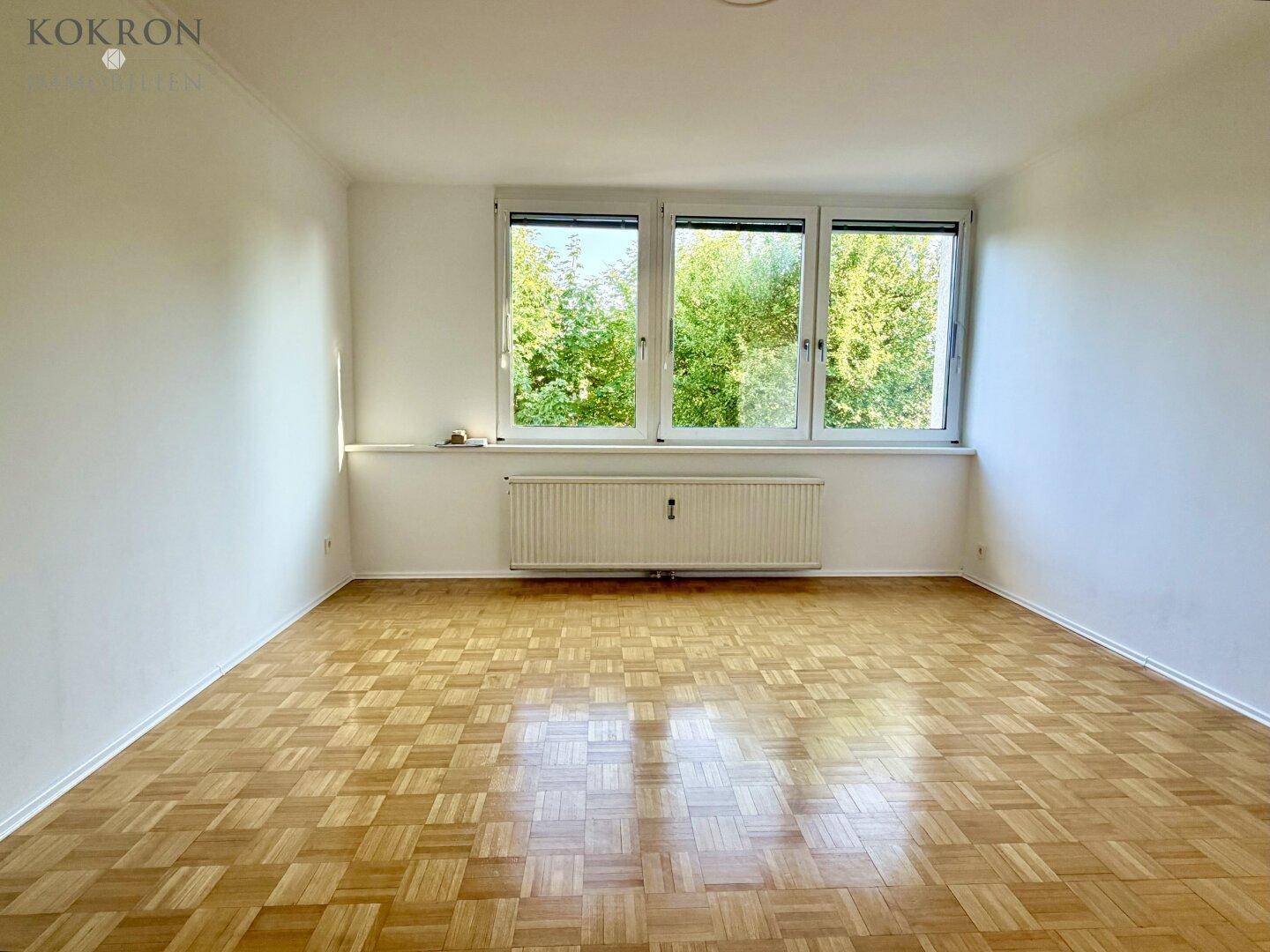 Leerer Raum mit Parkettboden und großem Fenster mit Blick auf grüne Bäume.