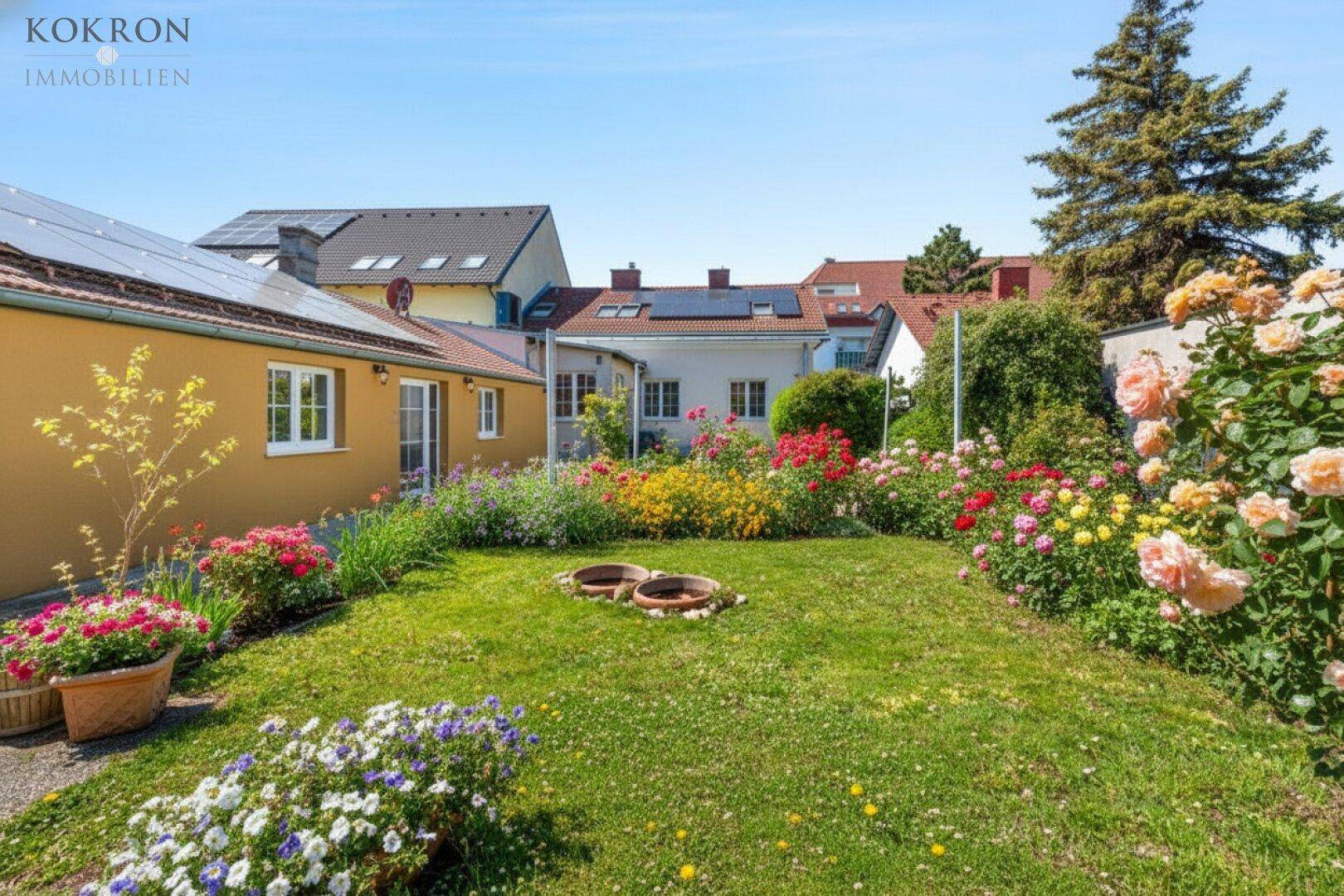 Gepflegter Garten mit Rasenfläche, bunten Blumenbeeten und Rosensträuchern unter blauem Himmel.