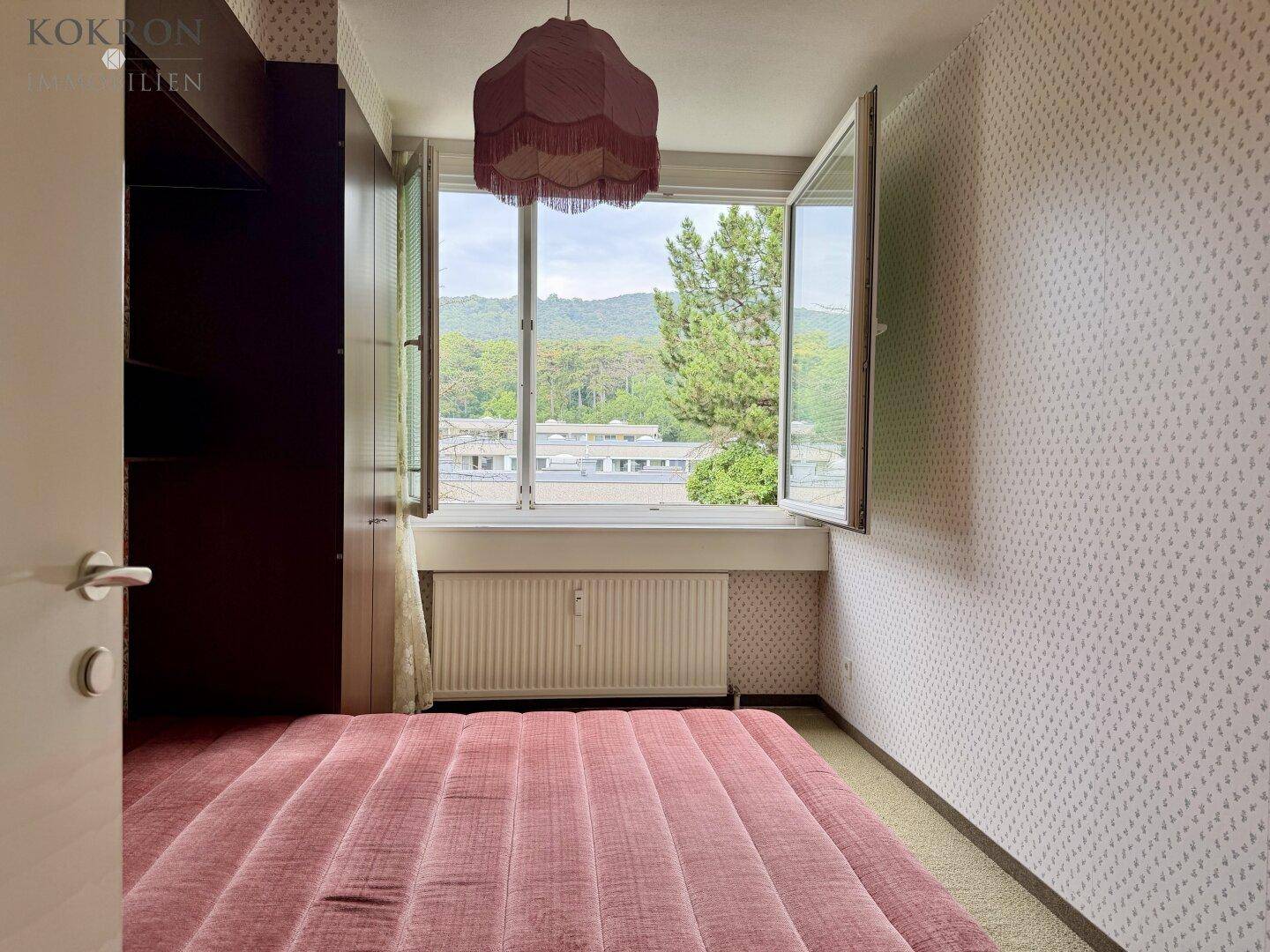 Schlafzimmer mit gemusterter Tapete, Fenster mit Blick ins Grüne und rosa Bett.