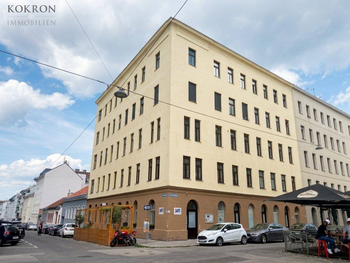 Mehrstöckiges Eckgebäude mit gelber Fassade und Geschäften im Erdgeschoss an einer belebten Straße.