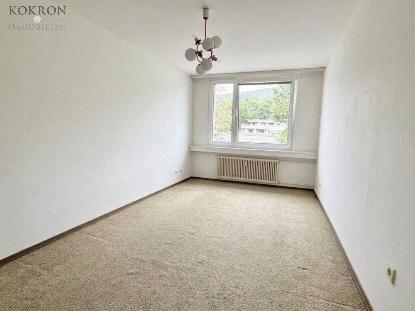 Heller Raum mit Teppichboden, großem Fenster und Heizkörper, bietet einen weiten Blick ins Grüne.