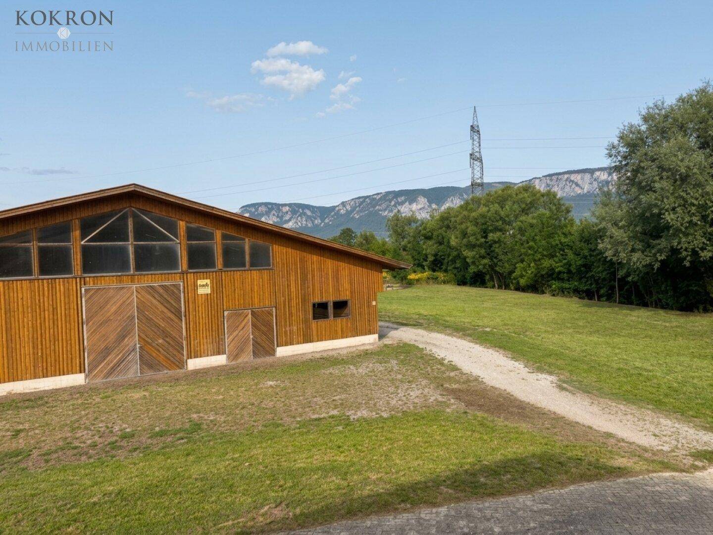 Holzgebäude in ländlicher Umgebung mit weitem Blick auf die Berge und grüne Felder unter blauem Himmel.