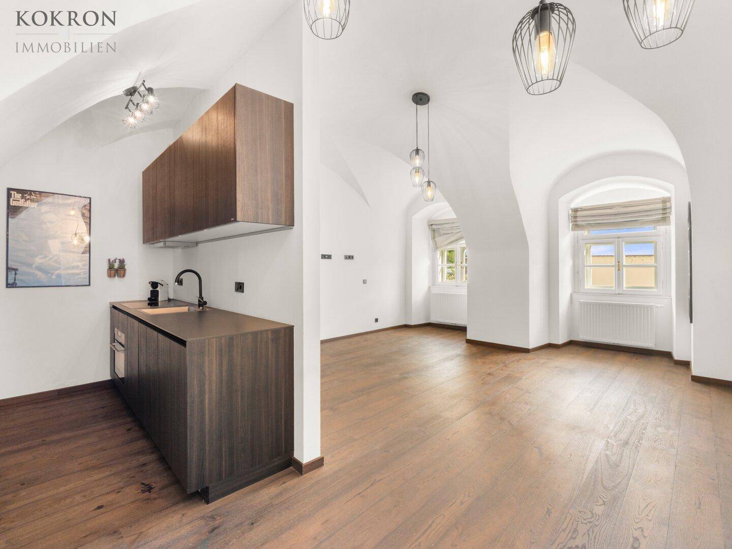 Geräumiger Wohnbereich mit integrierter Küchenzeile, Holzboden und gewölbter Decke in einem renovierten Gebäude.