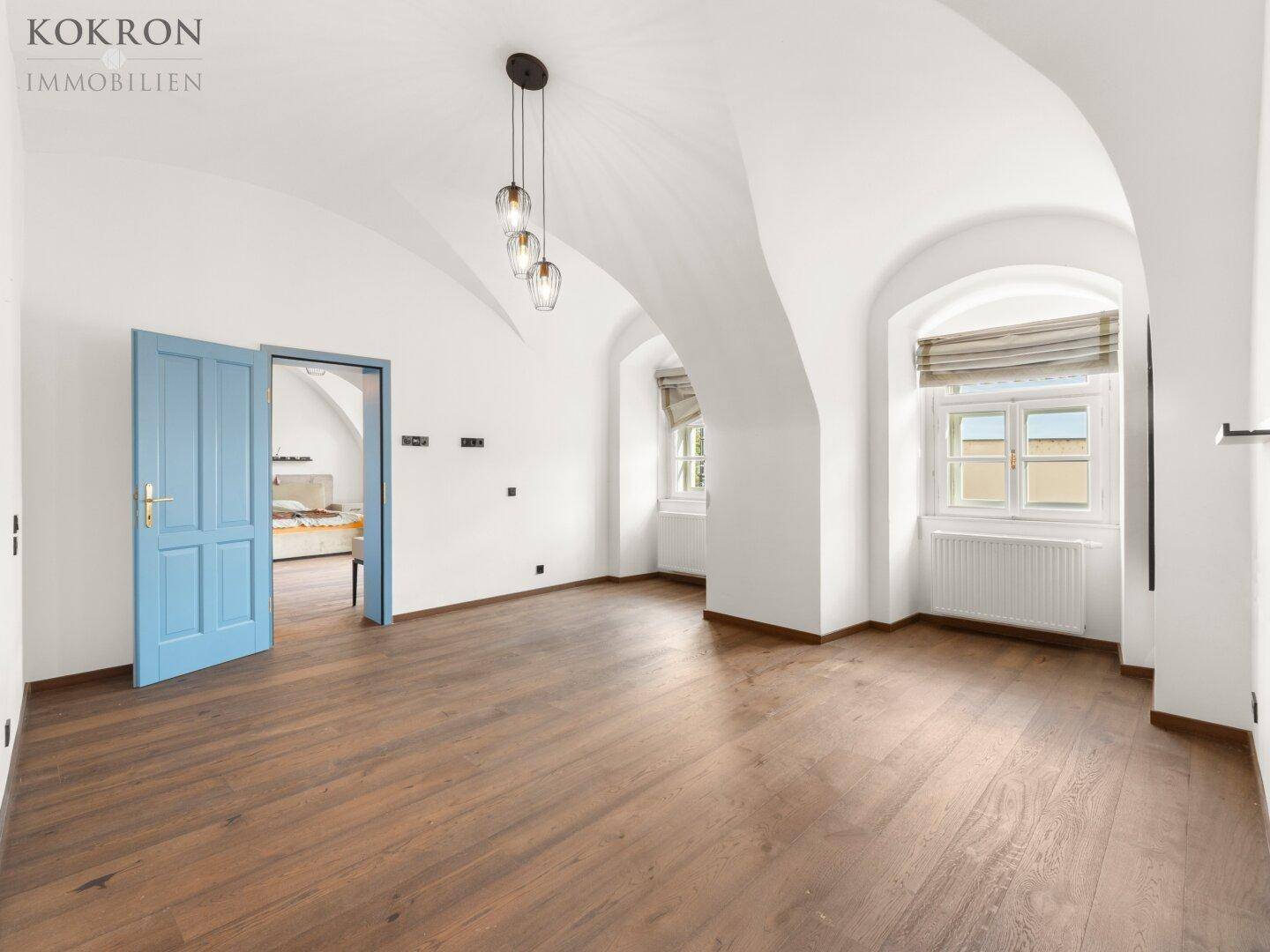 Heller Wohnbereich mit Holzboden, gewölbter Decke und einer auffälligen blauen Tür, die zu einem weiteren Raum führt.