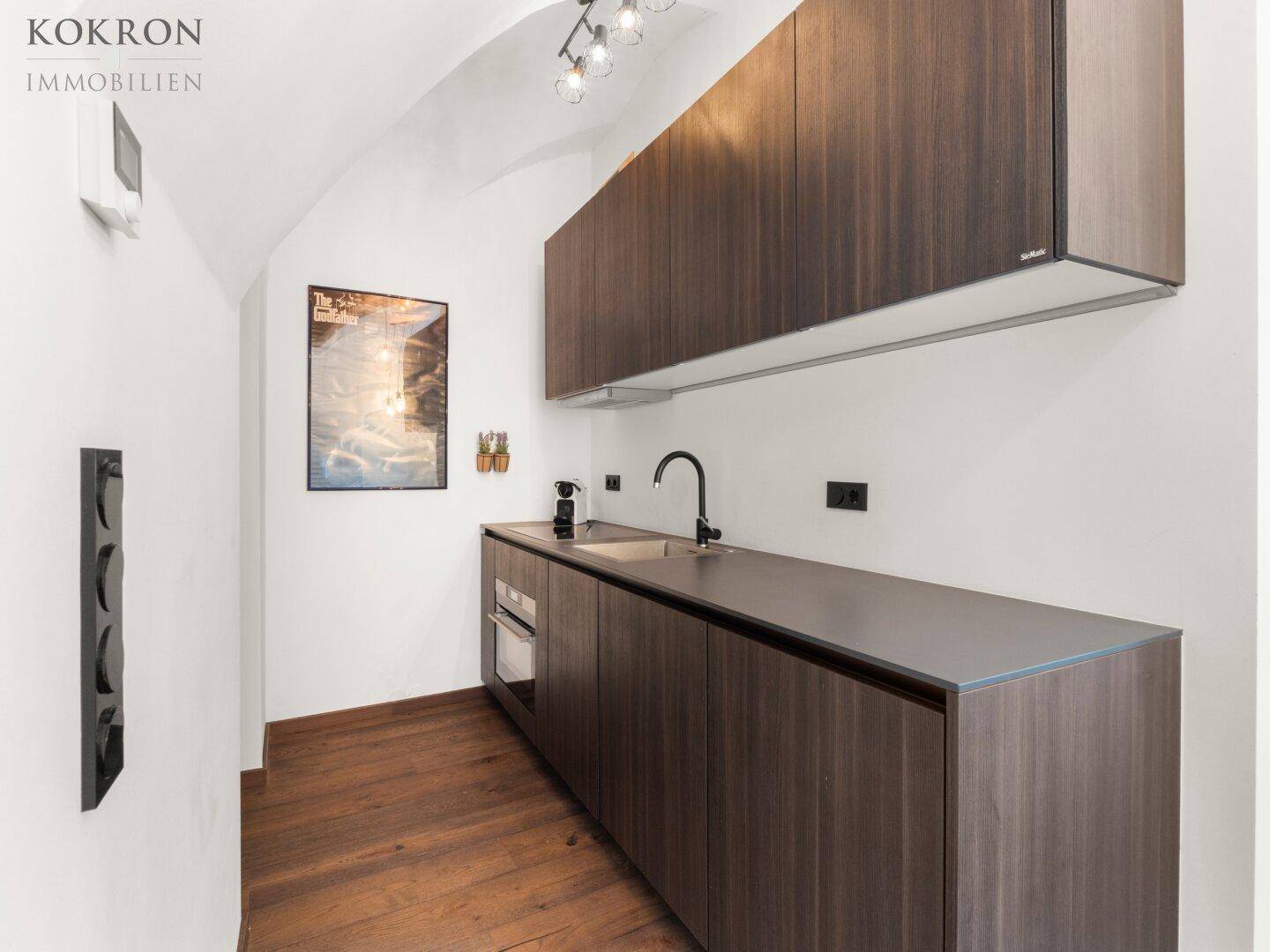 Moderne Küchenzeile mit dunklen Holzfronten, integriertem Backofen und minimalistischem Design in einem hellen Raum.
