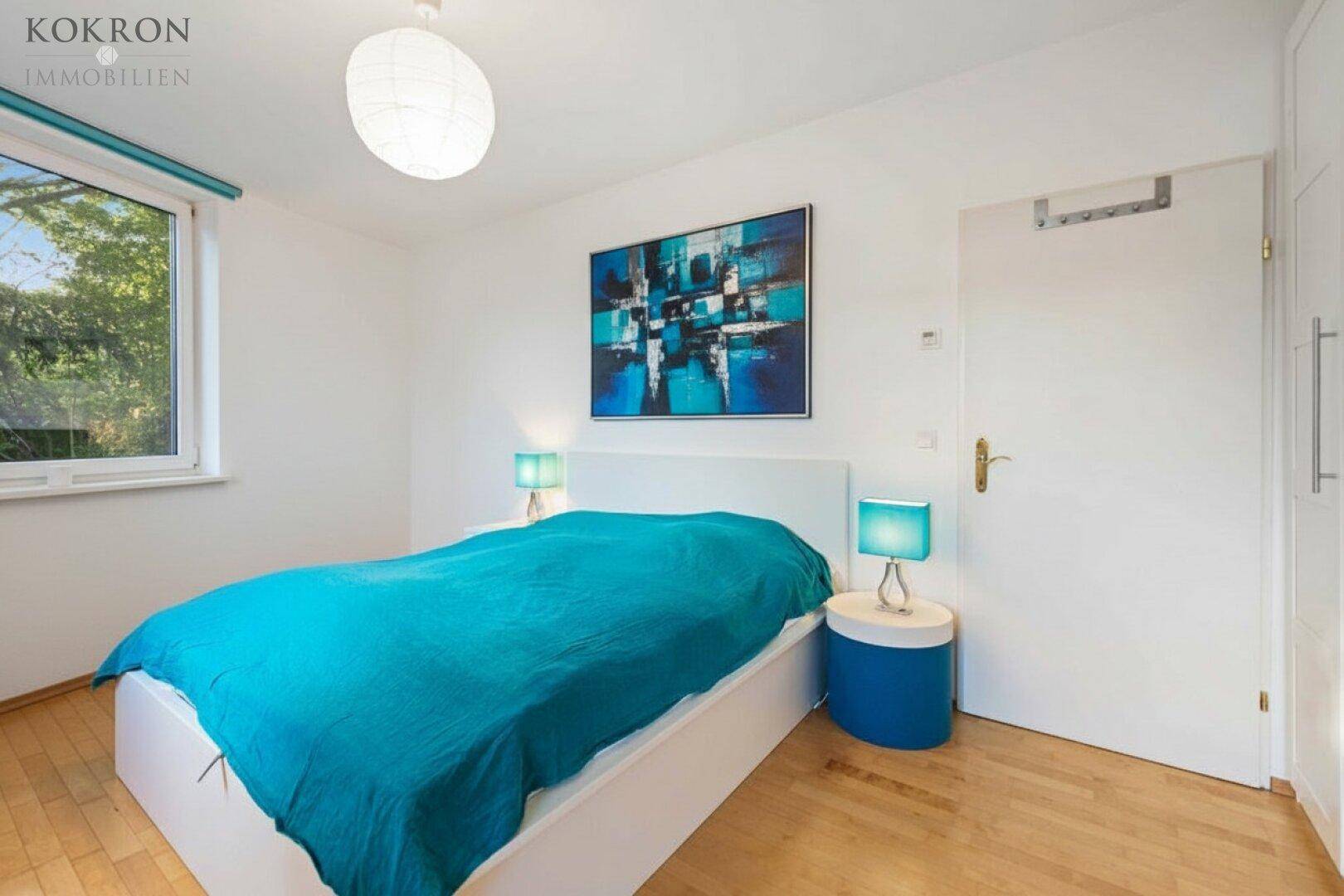 Helles Schlafzimmer mit Holzboden, großem Fenster und modernem Bett mit blauer Bettwäsche.