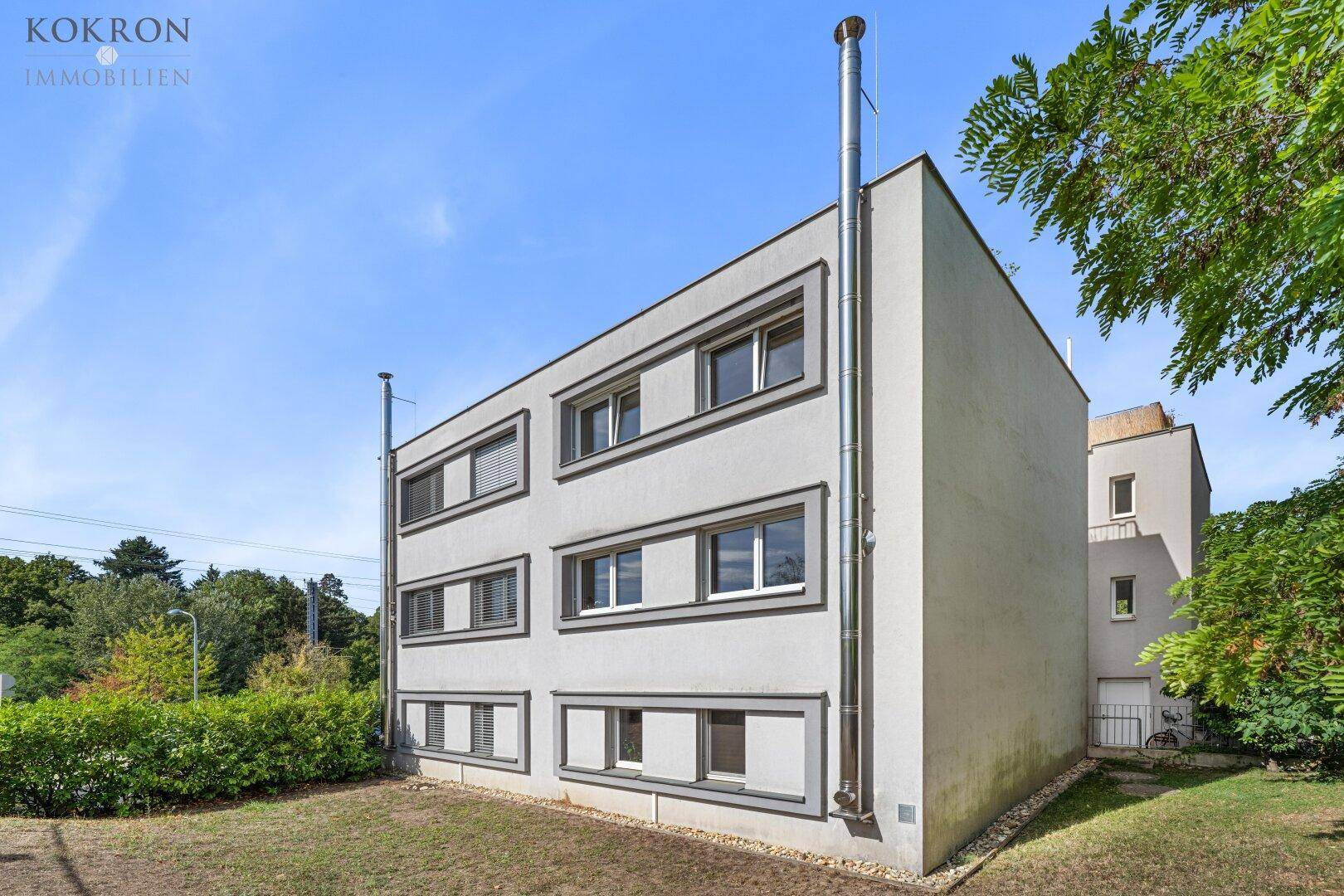 Moderne Gebäudefassade mit mehreren Fenstern und schlichtem Design, umgeben von Grünflächen.