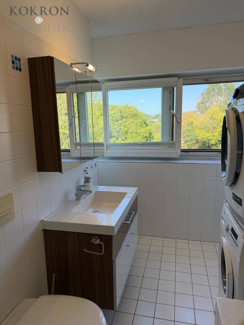 Funktionales Badezimmer mit Fenster, weißen Fliesen, Waschbecken und integrierter Waschmaschine und Trockner.