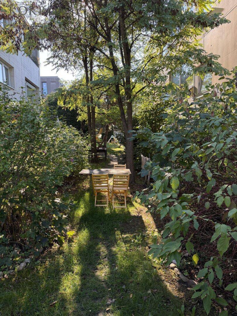 Idyllischer Gartenbereich mit Sitzgelegenheiten unter Bäumen, umgeben von üppigem Grün und Rasenfläche.