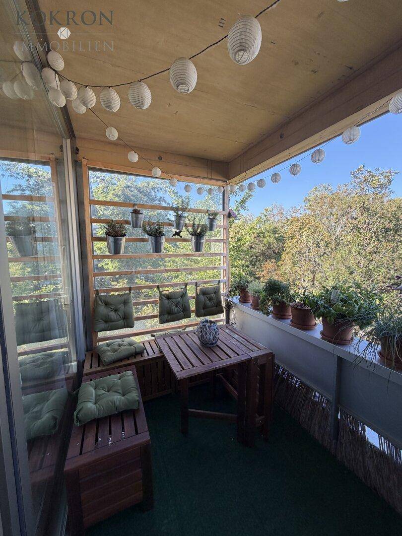 Gemütlicher Balkon mit Holztisch und Sitzgelegenheiten, umgeben von üppigem Grün und Lichterketten.