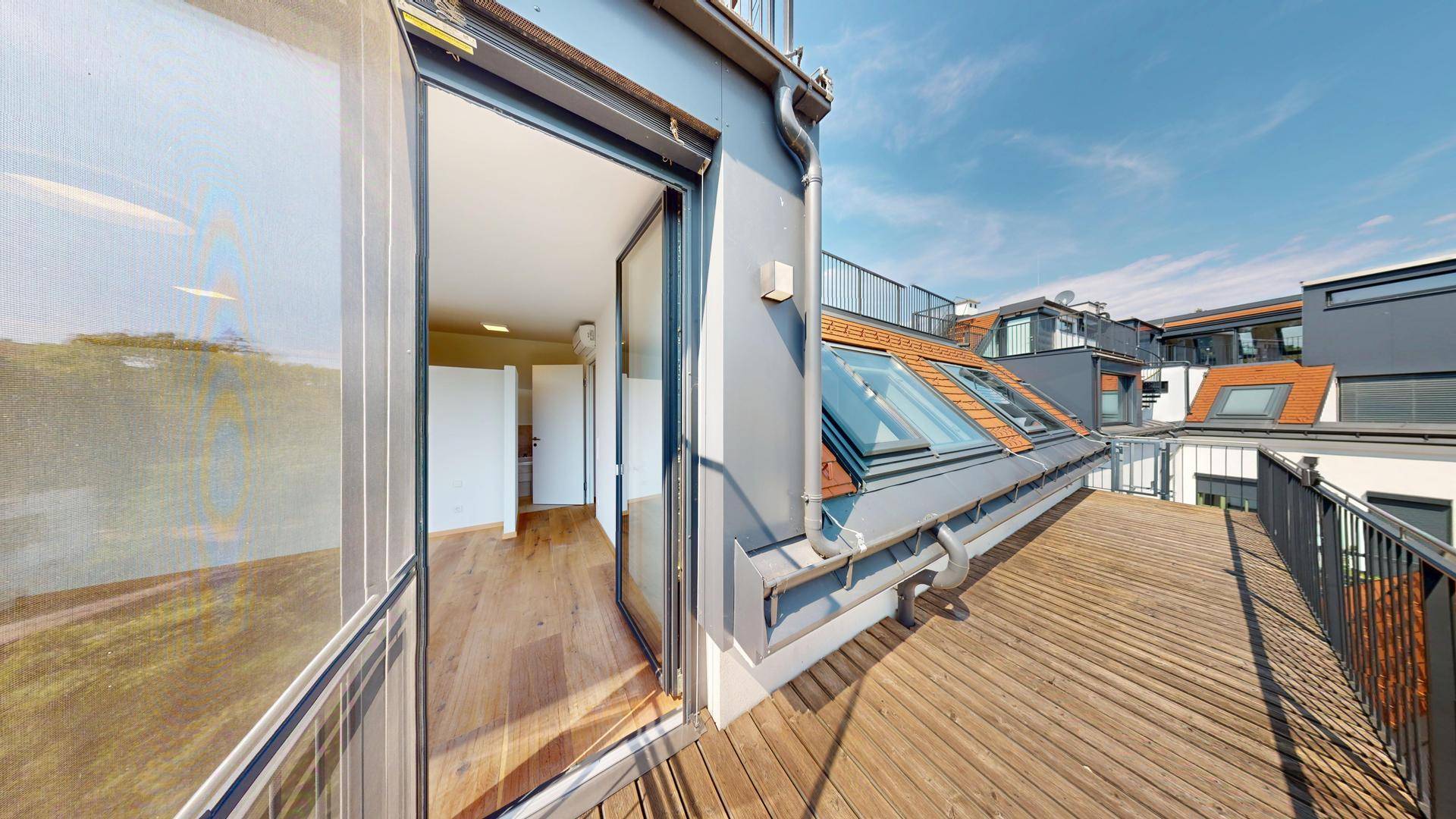 Geräumiger Holzbalkon mit Metallgeländer und Blick auf die Dächer und grüne Umgebung.