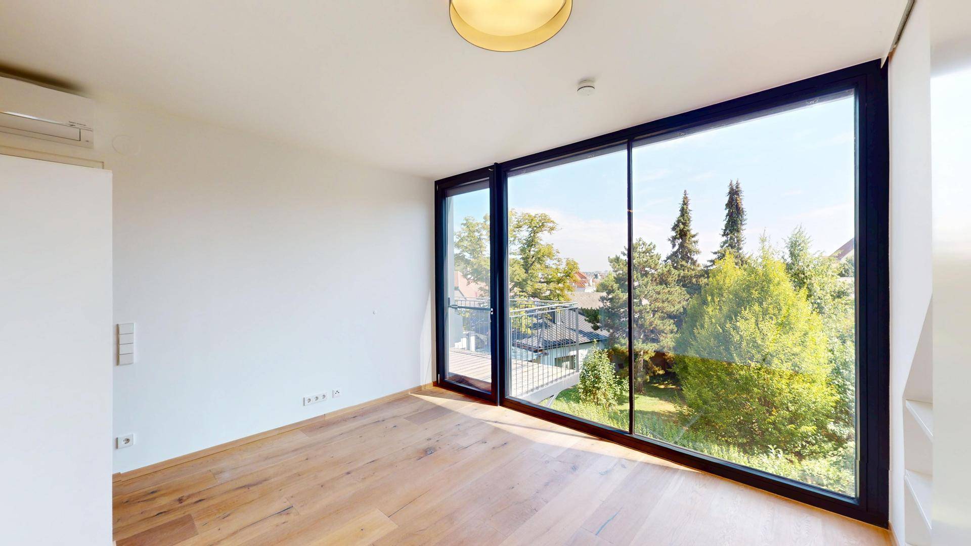 Heller Raum mit großen Fenstern, Holzboden und Zugang zu einem Balkon mit grüner Aussicht.