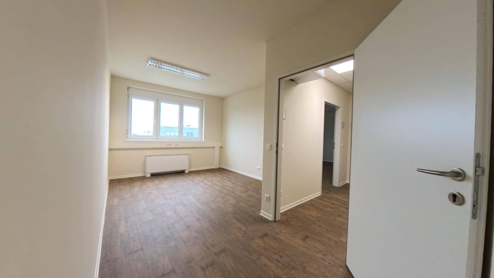 Helles Büro mit Holzboden und einer offen stehenden Tür, die den Zugang zu einem weiteren Raum ermöglicht.