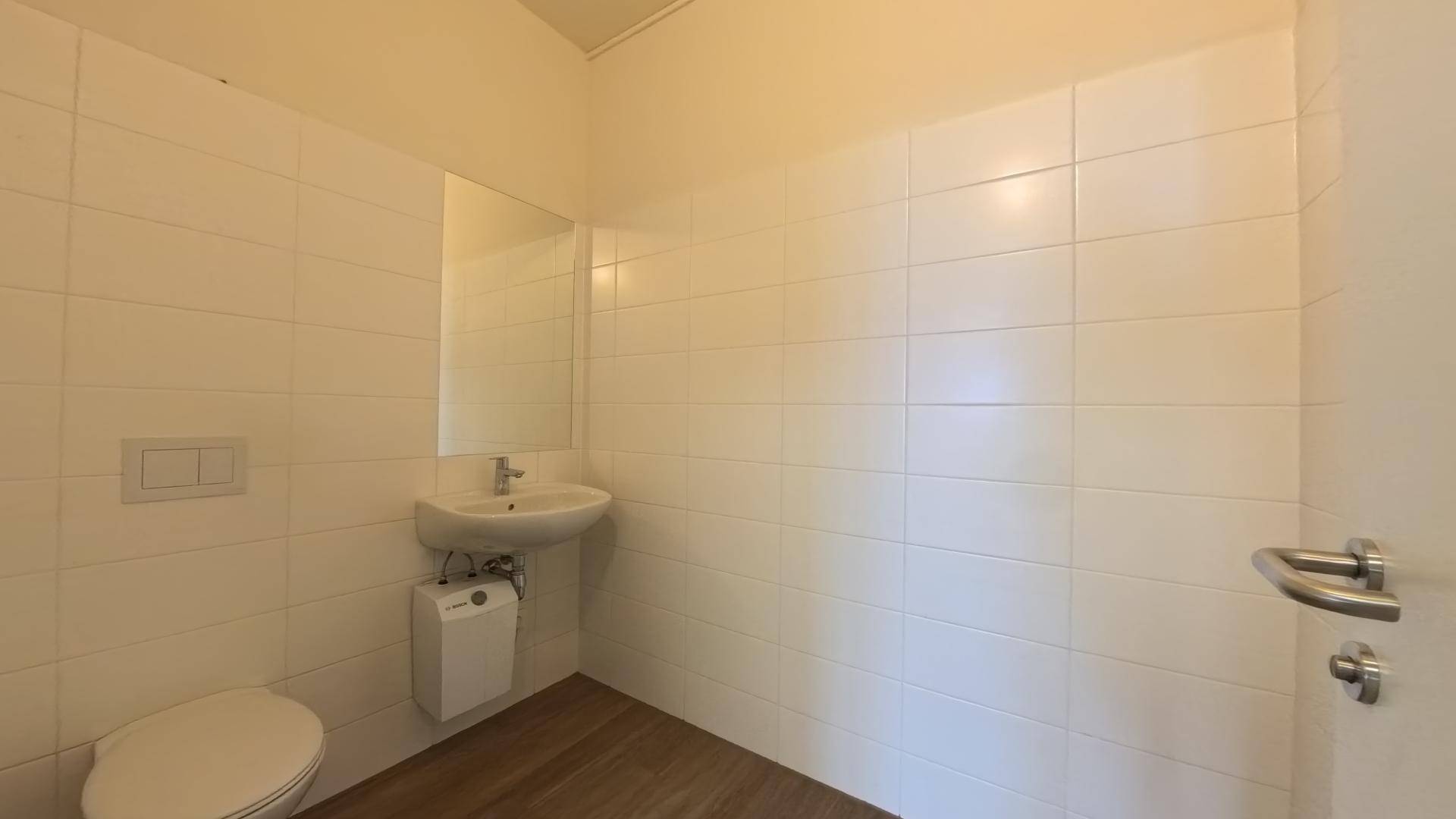 Helles Badezimmer mit weißen Fliesen, Waschbecken, Spiegel und wandhängender Toilette.