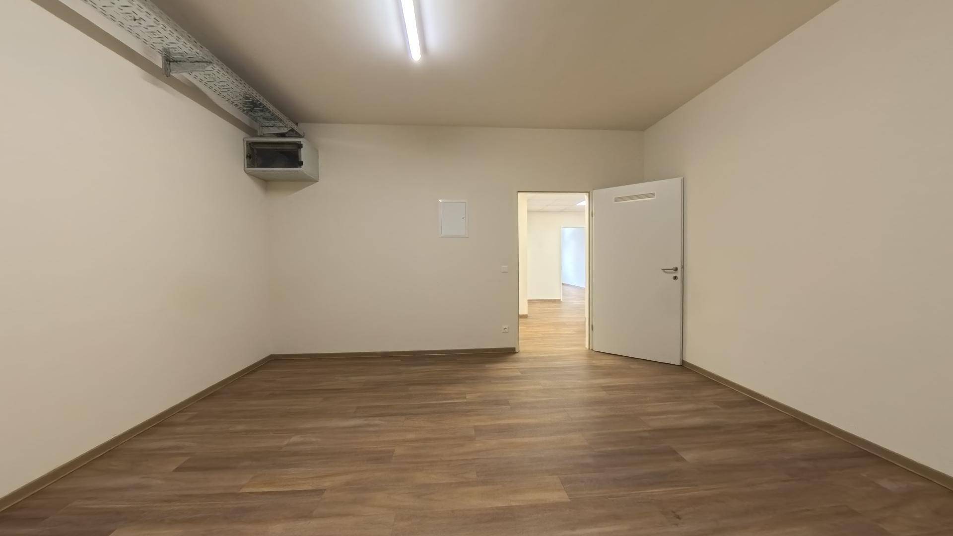 Büroraum mit Laminatboden, sichtbaren Lüftungsrohren und einer offenen Tür zu einem weiteren Bereich.
