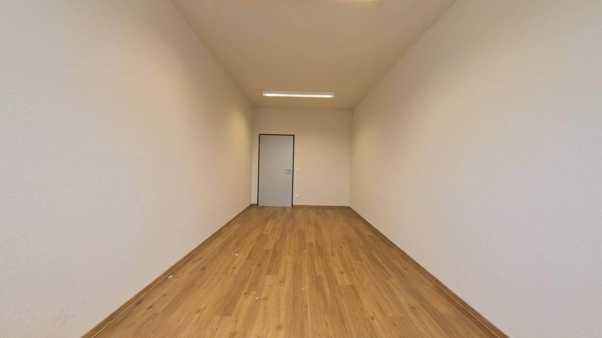 Leerer, langer Raum mit Laminatboden und weißen Wänden, der eine schlichte Tür an der Stirnseite zeigt.