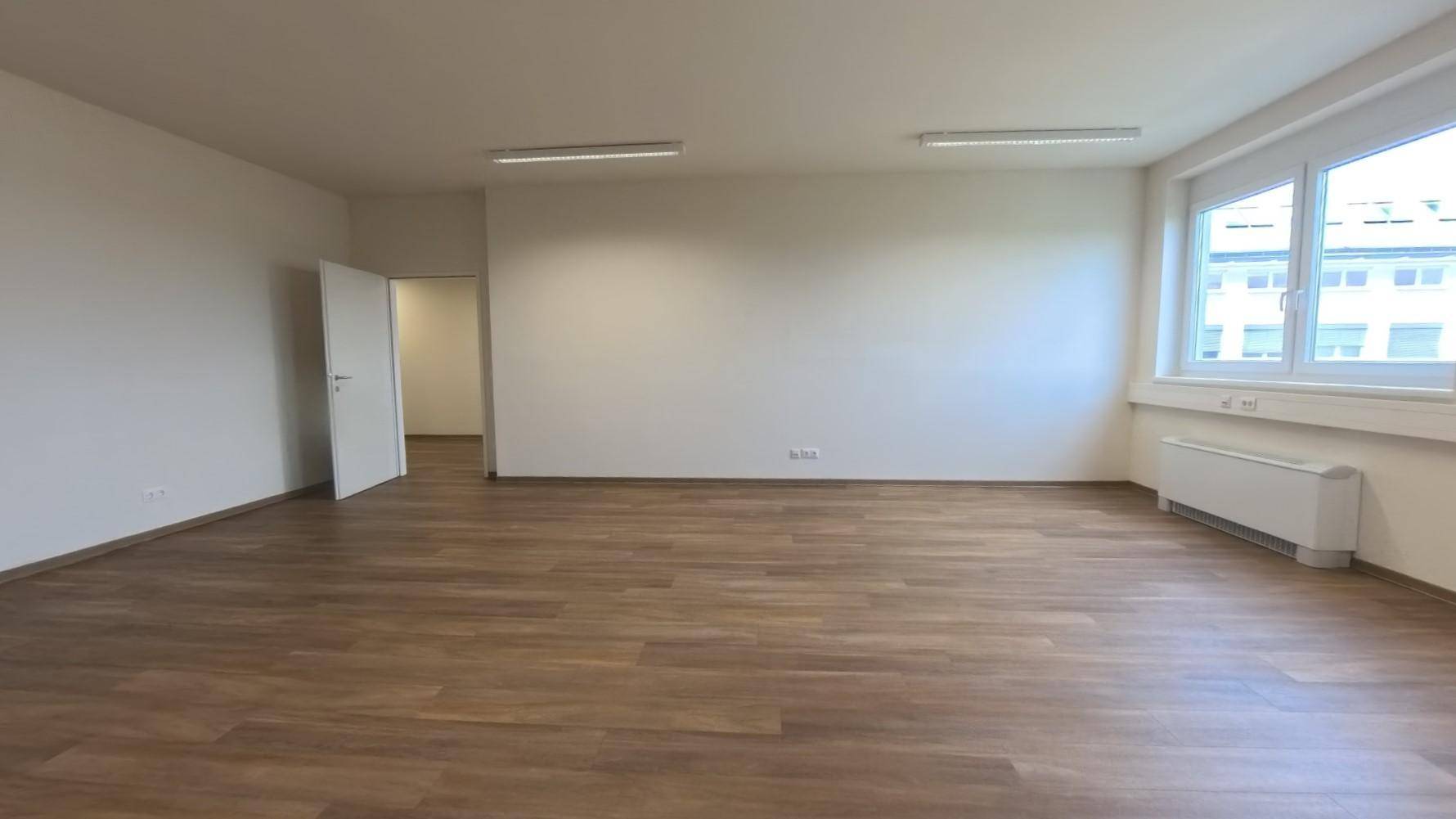 Heller Büroraum mit zwei Fenstern, Laminatboden und einer offenen Tür zu einem weiteren Bereich.