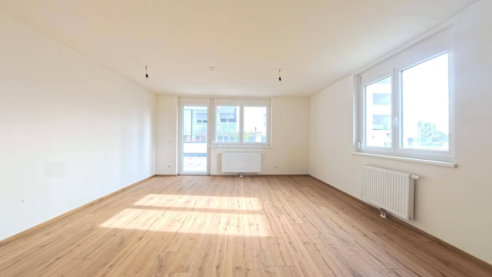 Helles, leeres Zimmer mit Laminatboden und Zugang zu einem Balkon oder einer Terrasse.
