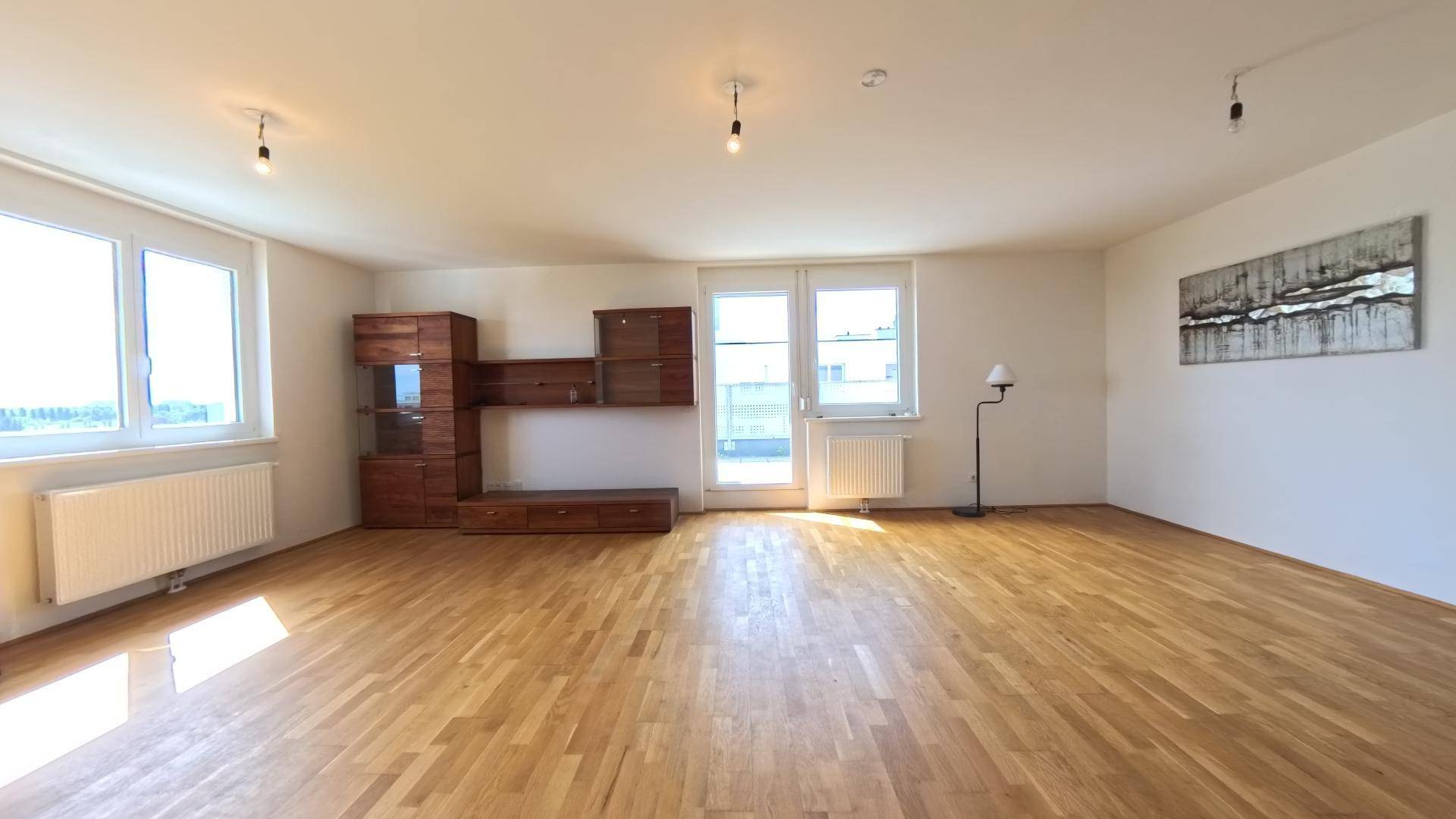 Geräumiges Wohnzimmer mit hellem Holzparkett, großen Fenstern und Zugang zu einem Außenbereich.