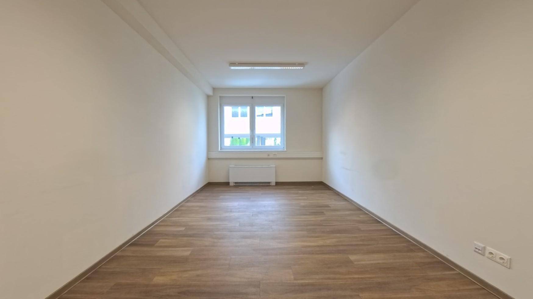 Leerer Büroraum mit Laminatboden, einem Fenster und neutralen Wänden, bereit zur Einrichtung.