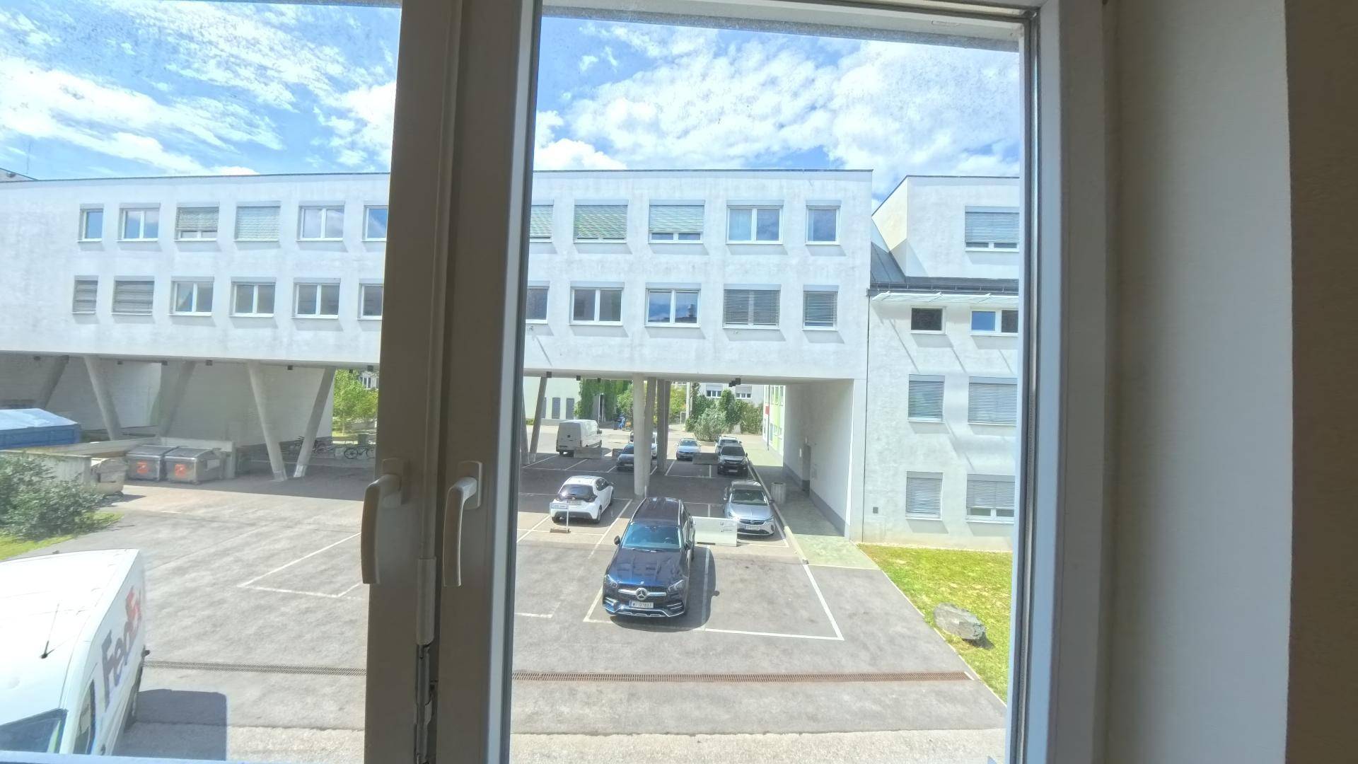 Blick aus dem Fenster auf einen Parkplatz und ein modernes Bürogebäude mit vielen Fenstern.