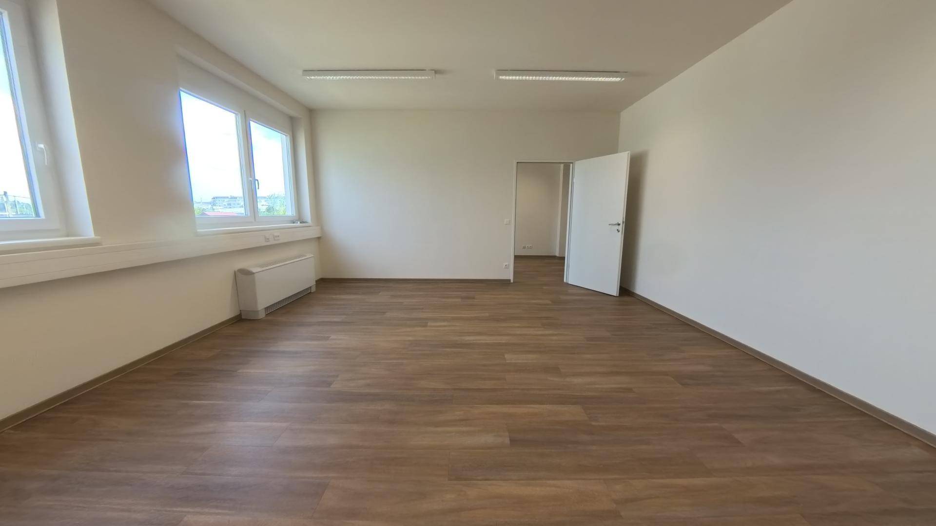 Heller Büroraum mit zwei Fenstern, Laminatboden und einer offenen Tür zu einem weiteren Bereich.