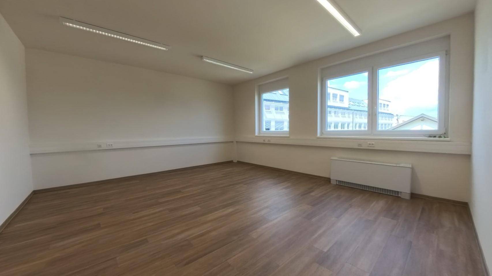 Heller Büroraum mit zwei Fenstern, Laminatboden und schlichten, hellen Wänden, ideal für Arbeitsplätze.