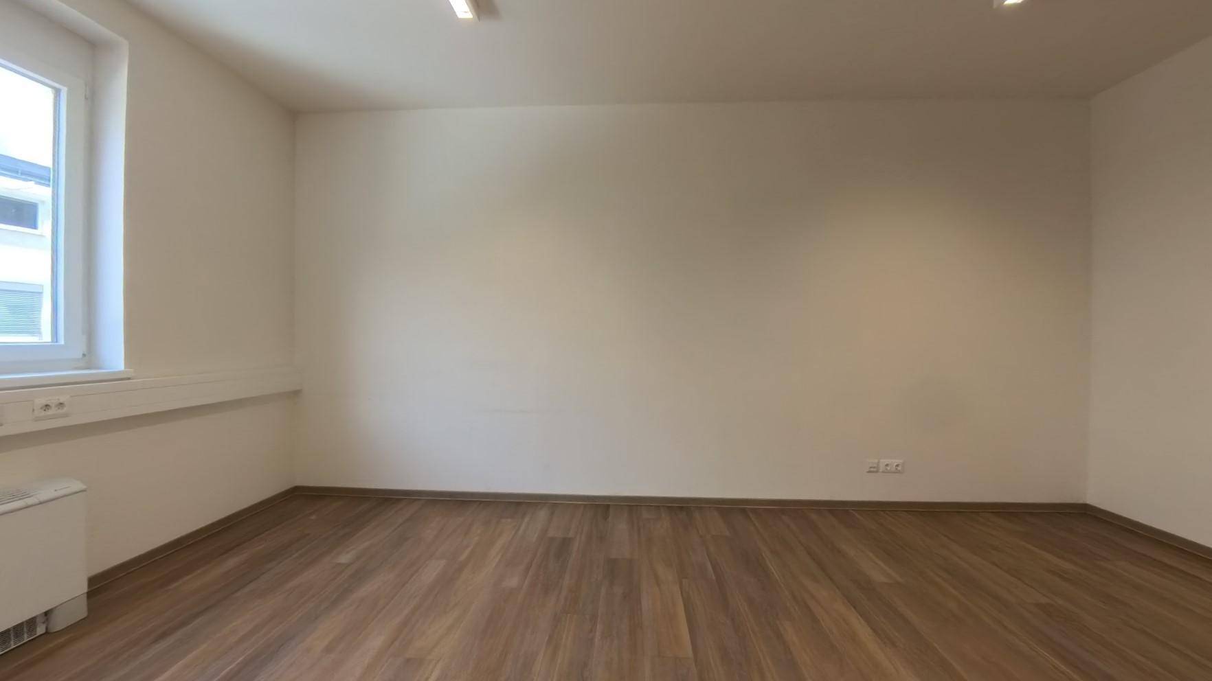 Heller Büroraum mit großem Fenster, Laminatboden und schlichten, hellen Wänden, ideal für Arbeitsplätze.