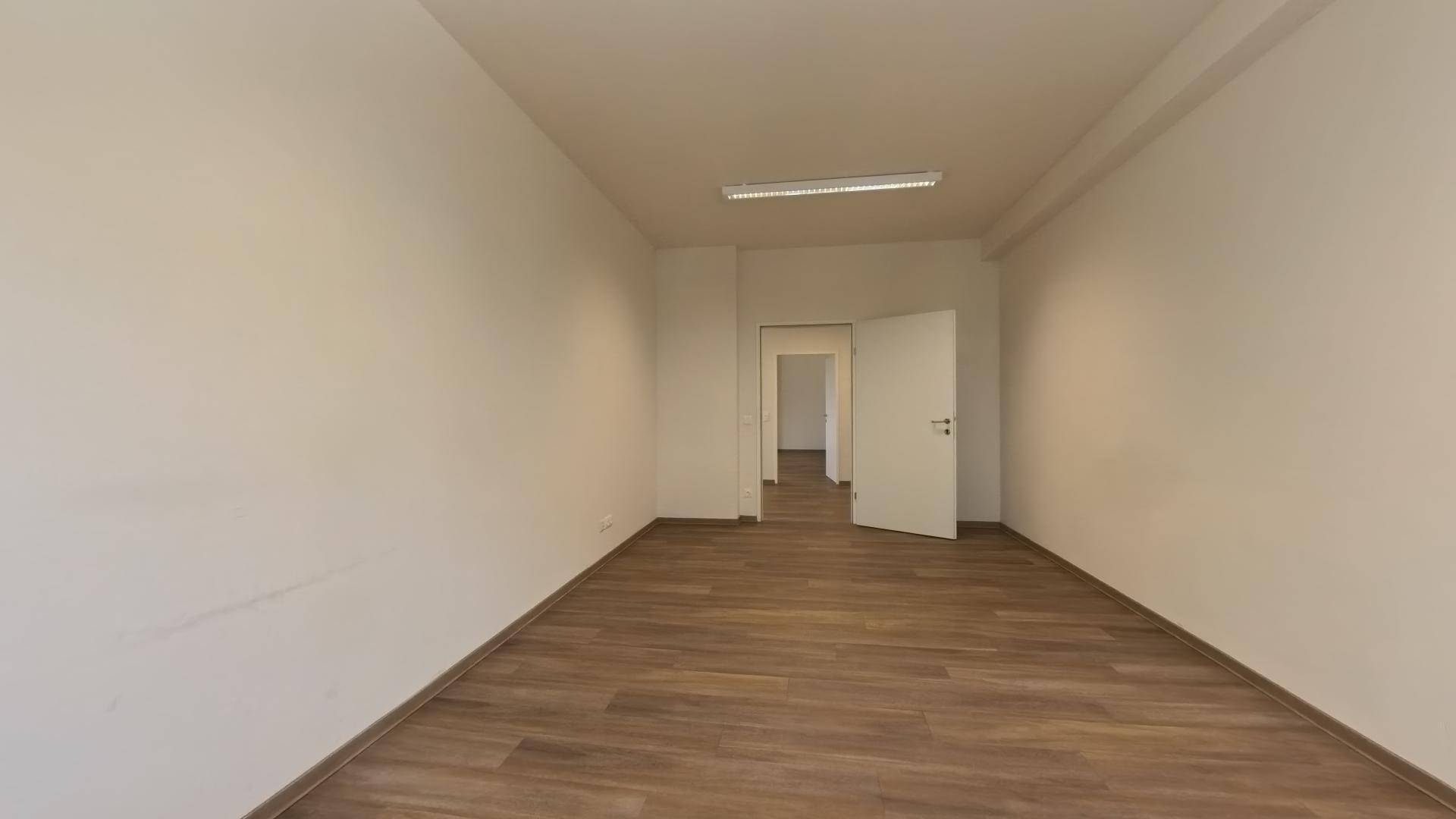 Geräumiger, leerer Büroraum mit Laminatboden und einer offenen Tür zu einem weiteren Bereich.